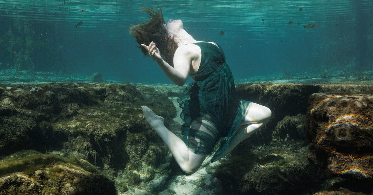 A woman in a black dress is underwater, kneeling on rocks, with her head tilted back and hair flowing.