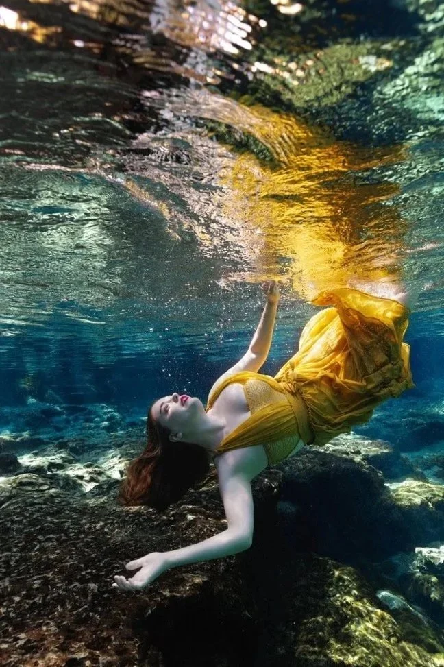 A woman in a yellow dress floating underwater near rocks, with her eyes closed and one arm raised towards the water's surface.