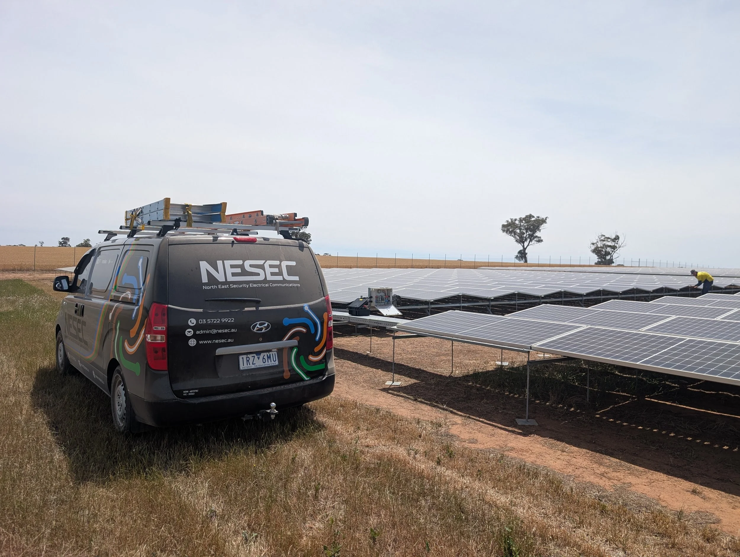 A security van labeled NSSEC parked beside solar panels in a rural area with open fields and a few trees.
