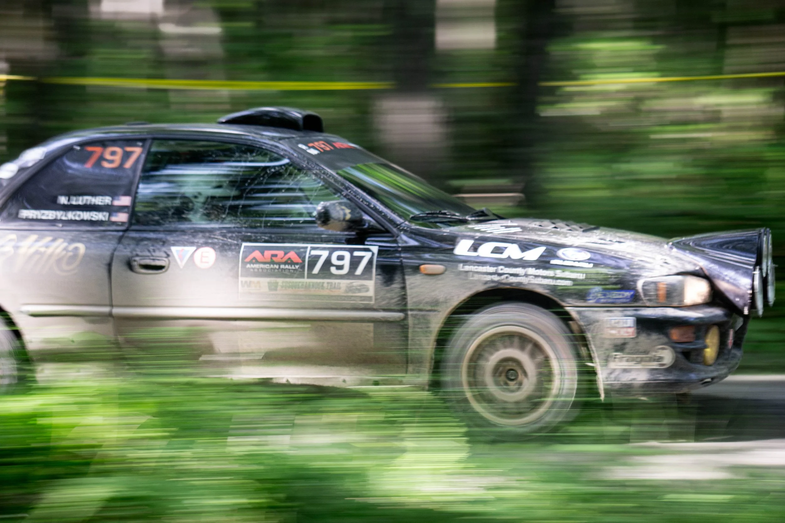 A black race car speeding through a wooded area, with a blurred background indicating motion. The car has red and white logos and the number 97 on the side.