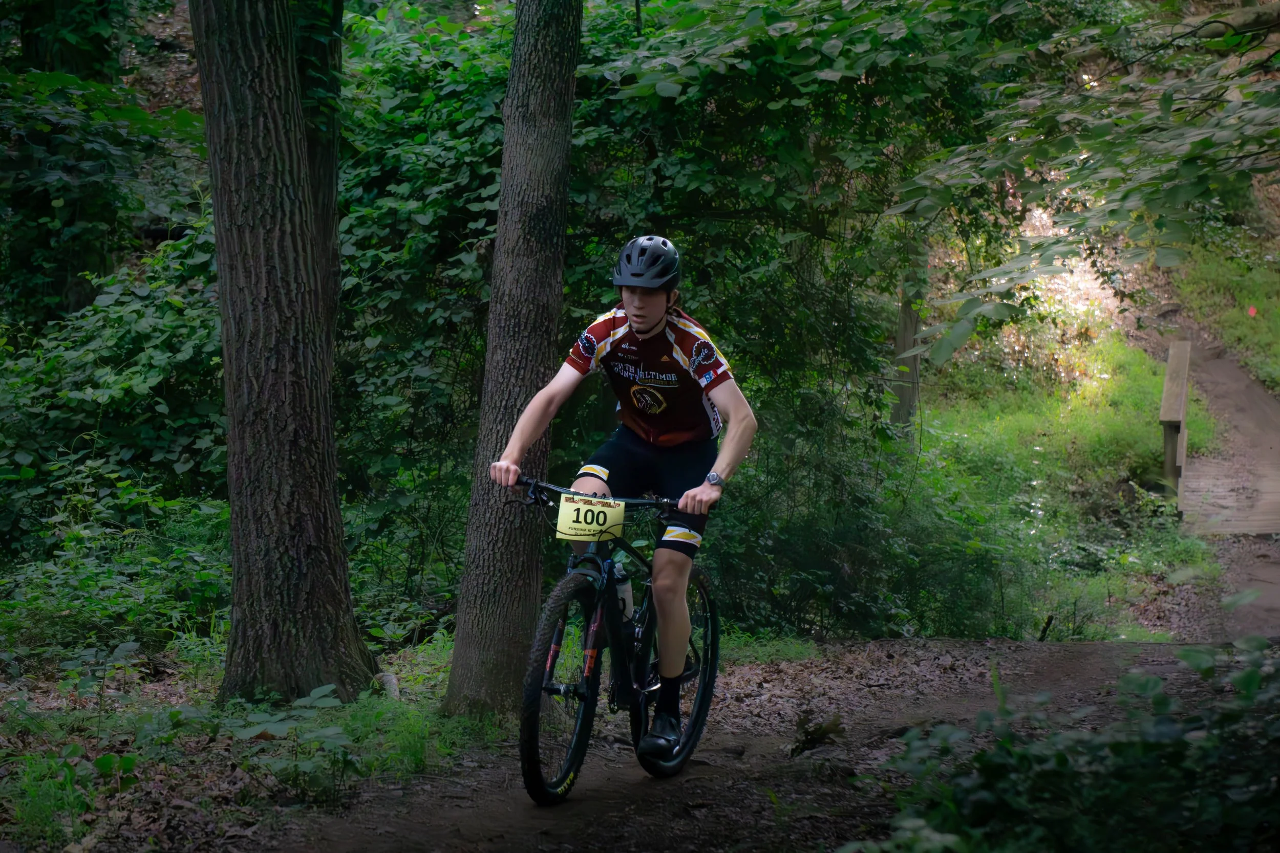 A young male cyclist wearing a helmet and a colorful cycling jersey riding a mountain bike on a forest trail surrounded by trees and green foliage.