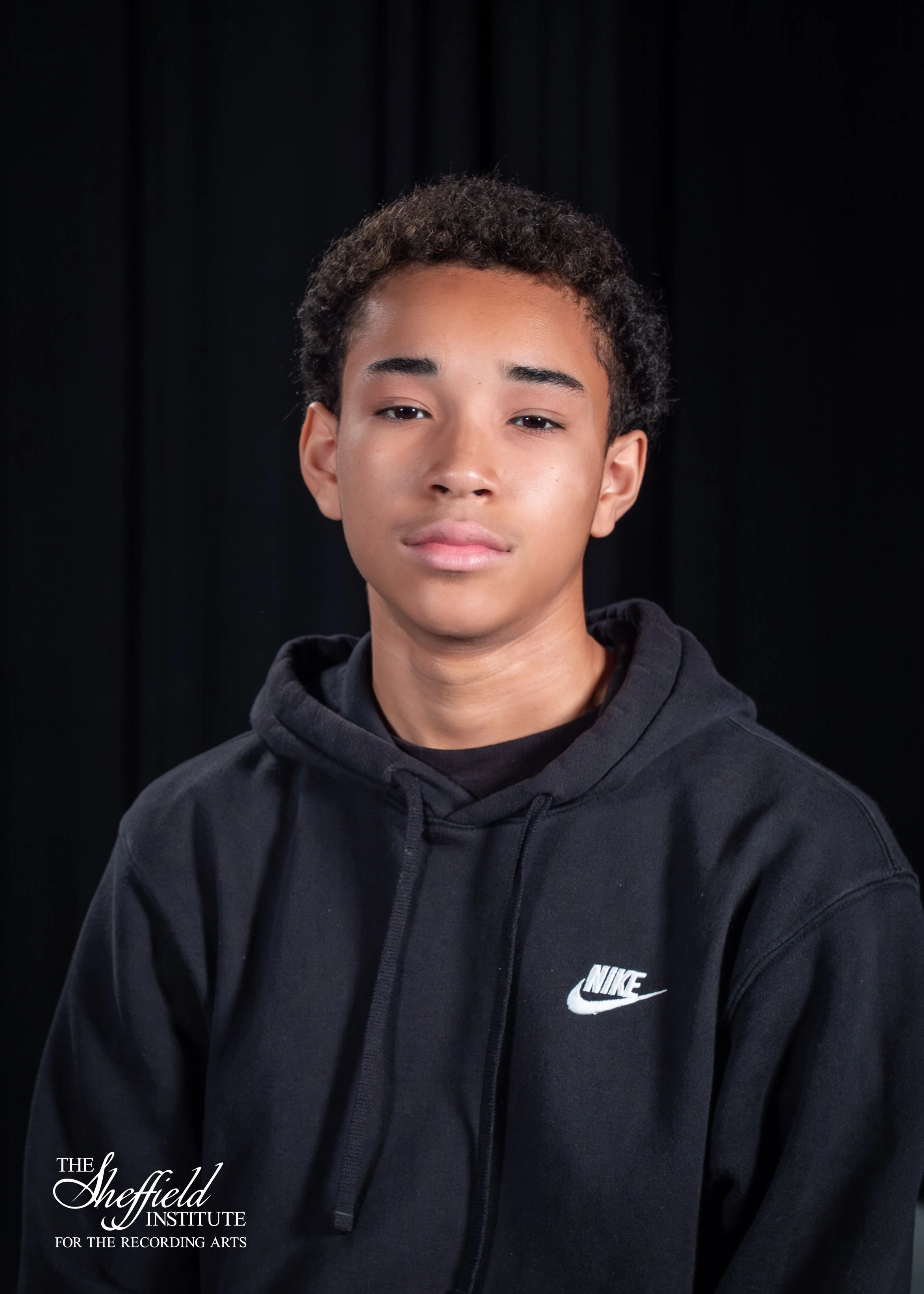 Young person with short curly hair wearing a black Nike hoodie, posing against a dark background.
