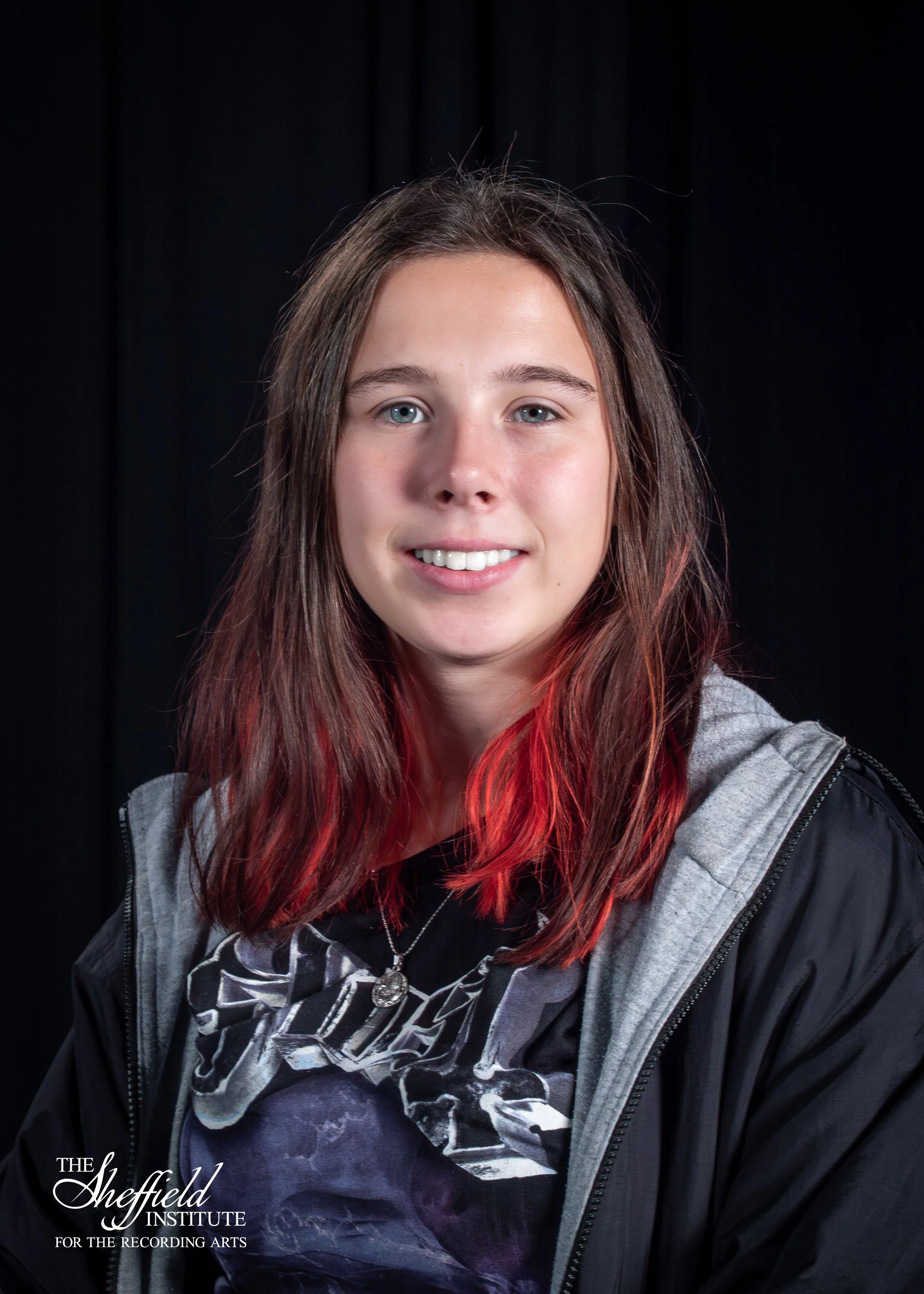 Young woman with blue eyes and long brown hair with red tips, wearing a black and gray jacket and a graphic t-shirt, posing against a black background, with the logo of The Sheffield Institute for the Recording Arts in the lower left corner.