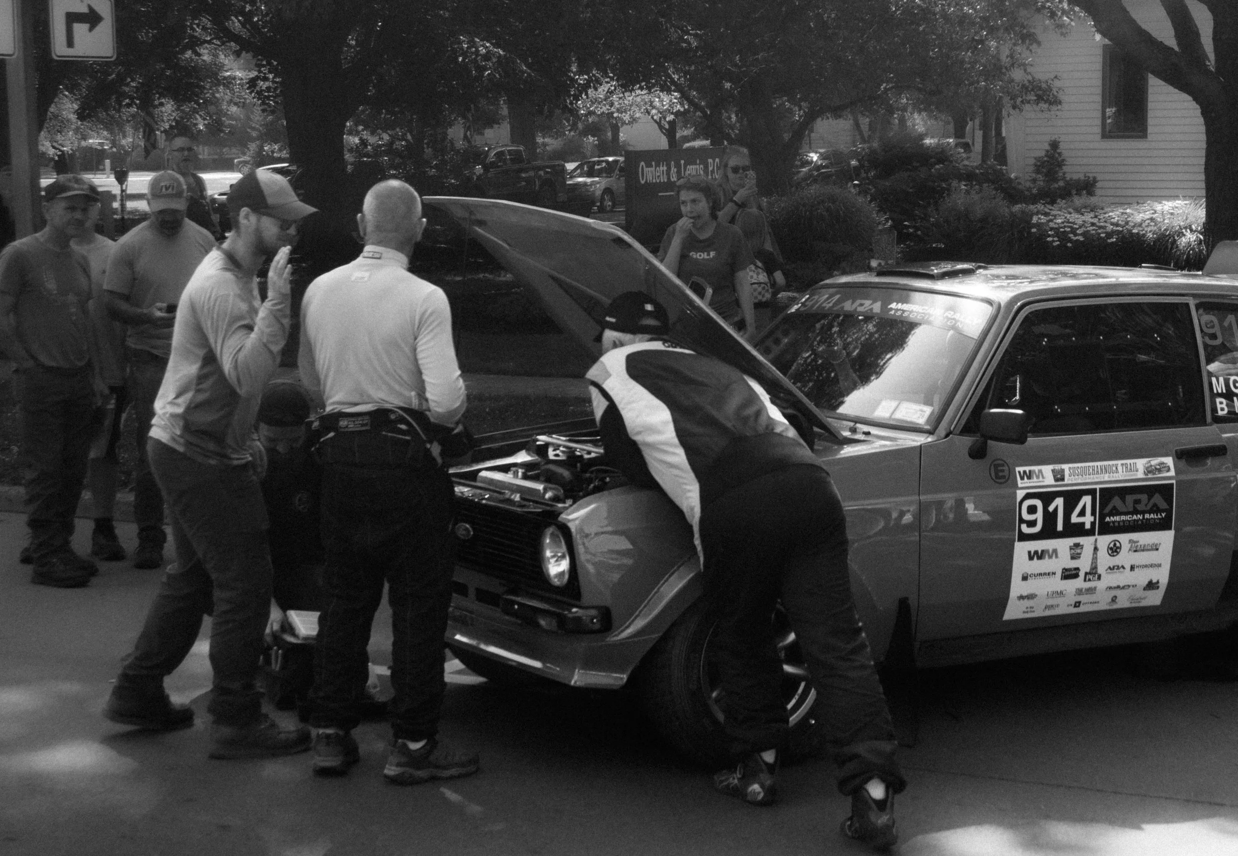 A group of people gathered around a car at an outdoor event, with trees and buildings in the background.