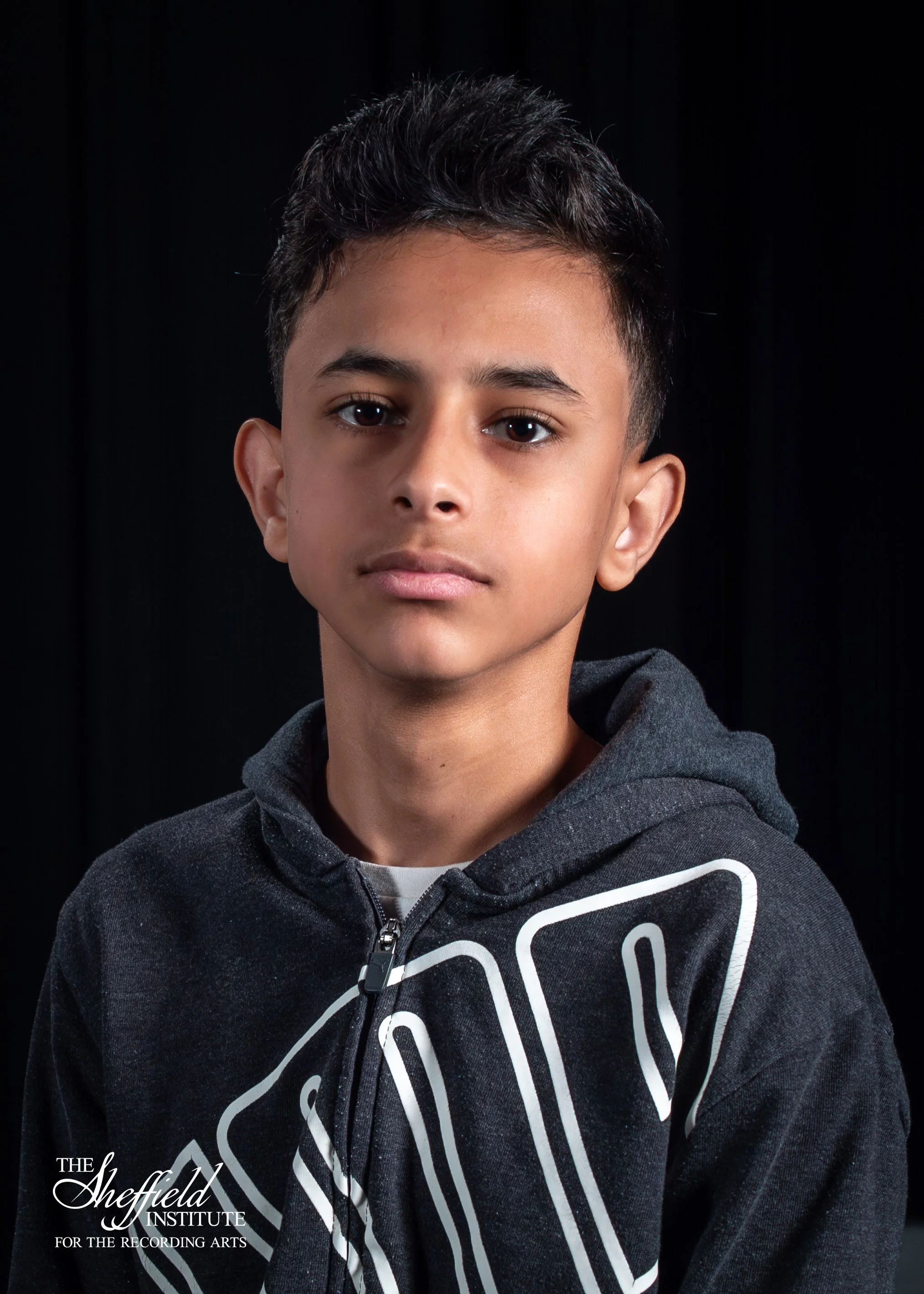 A young boy with short dark hair and light brown skin, wearing a black hoodie with white lettering, stands against a black background at The Sheffield Institute for the Recording Arts.