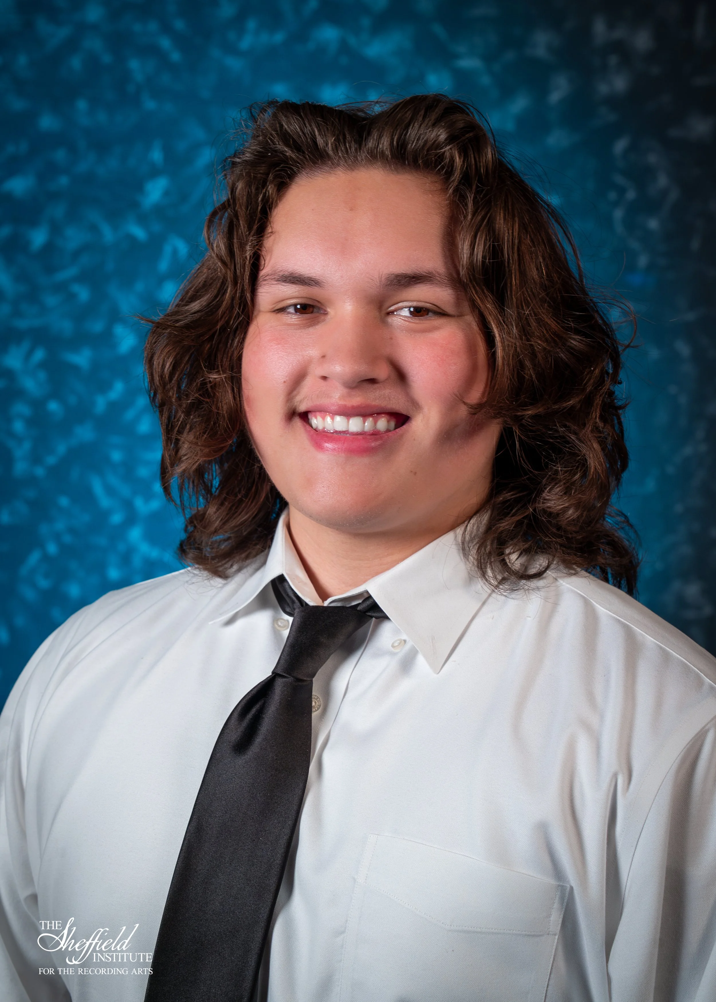 A young man with shoulder-length wavy brown hair, wearing a white dress shirt and black tie, smiling against a textured blue background. The Sheffield Institute logo is visible in the bottom left corner.