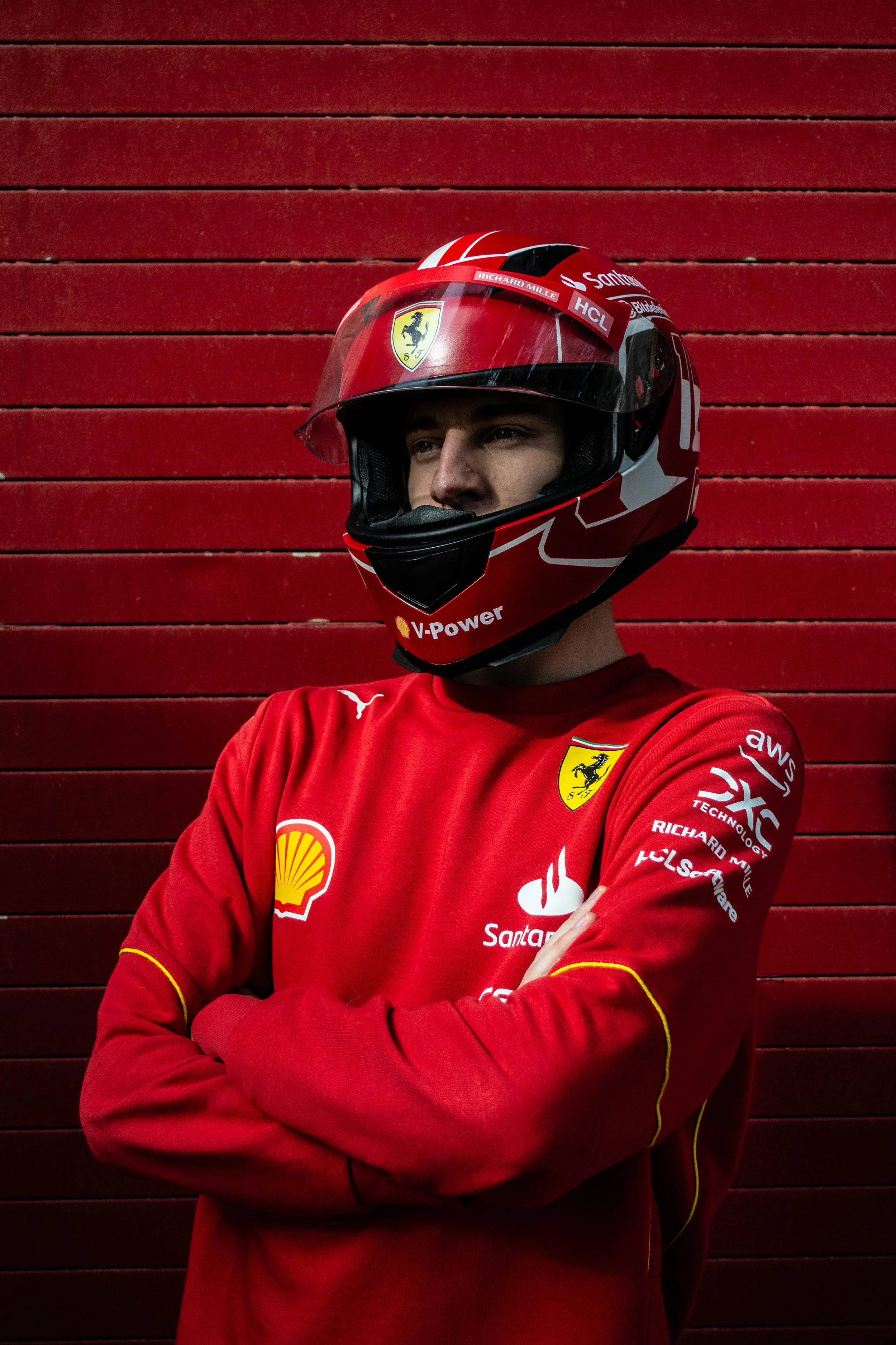A person wearing a red Ferrari racing helmet and a red racing jacket with sponsors and Ferrari logos stands against a red wooden wall with their arms crossed.