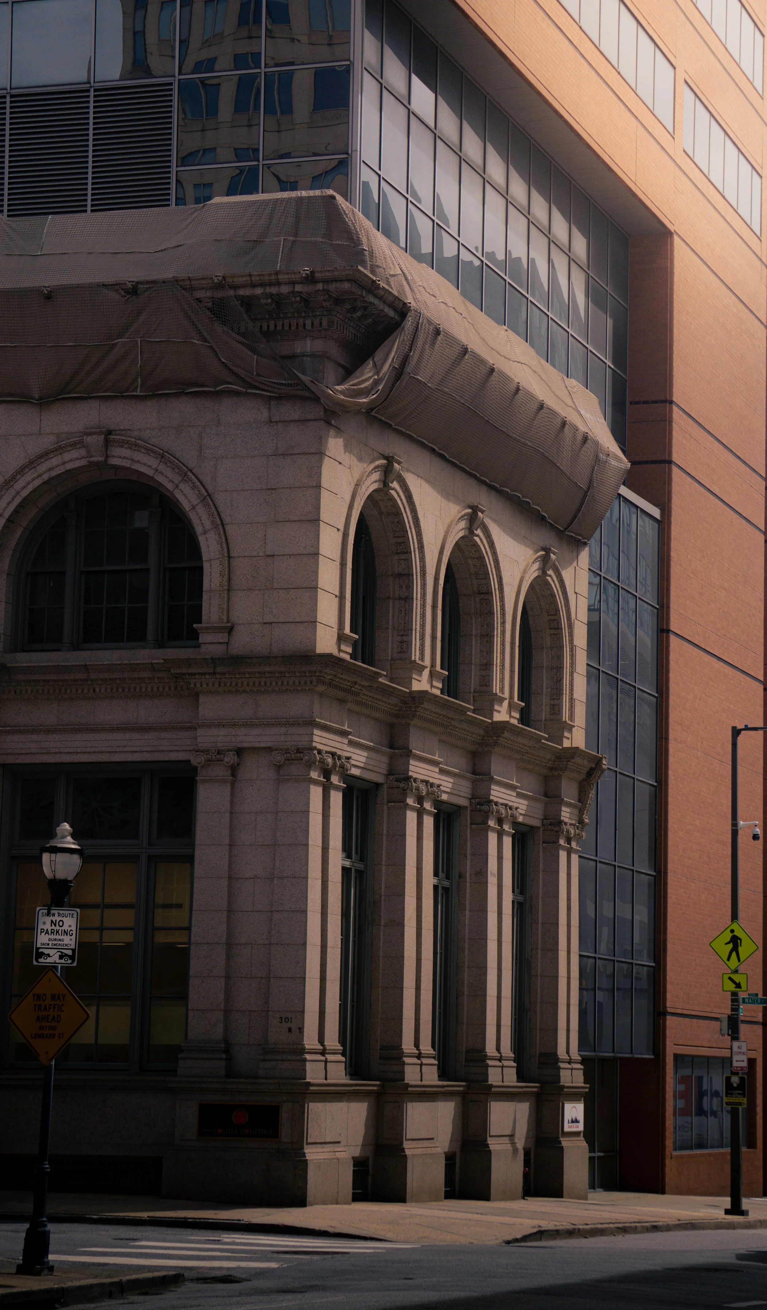 A historic building with arched windows and stone architecture, partially covered by construction fencing or netting, adjacent to a modern glass and brick building in an urban setting. Street signs and a pedestrian crossing sign are visible in the fo