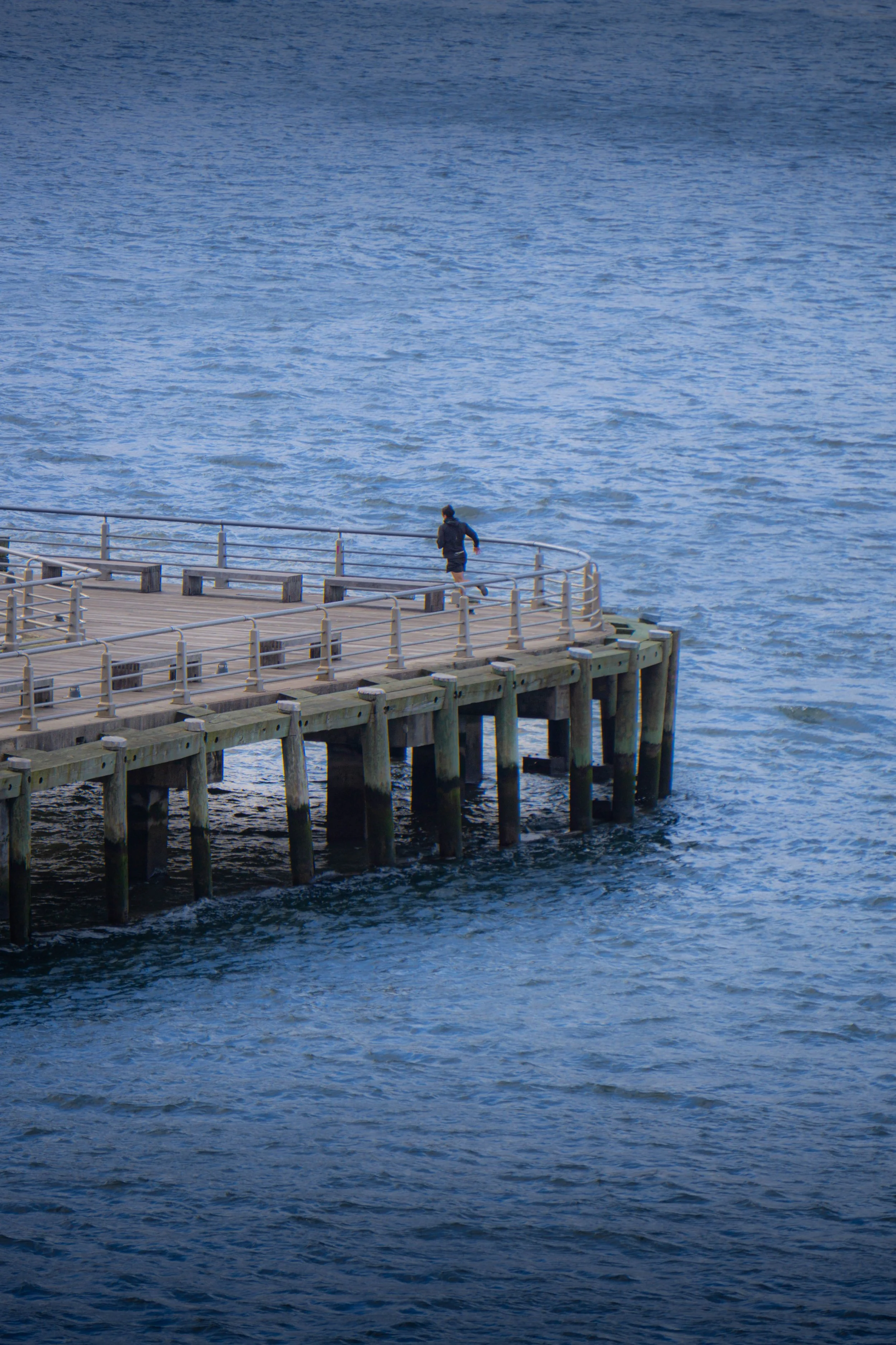 A person running on a wooden pier over the ocean with gentle waves and a clear blue sky.