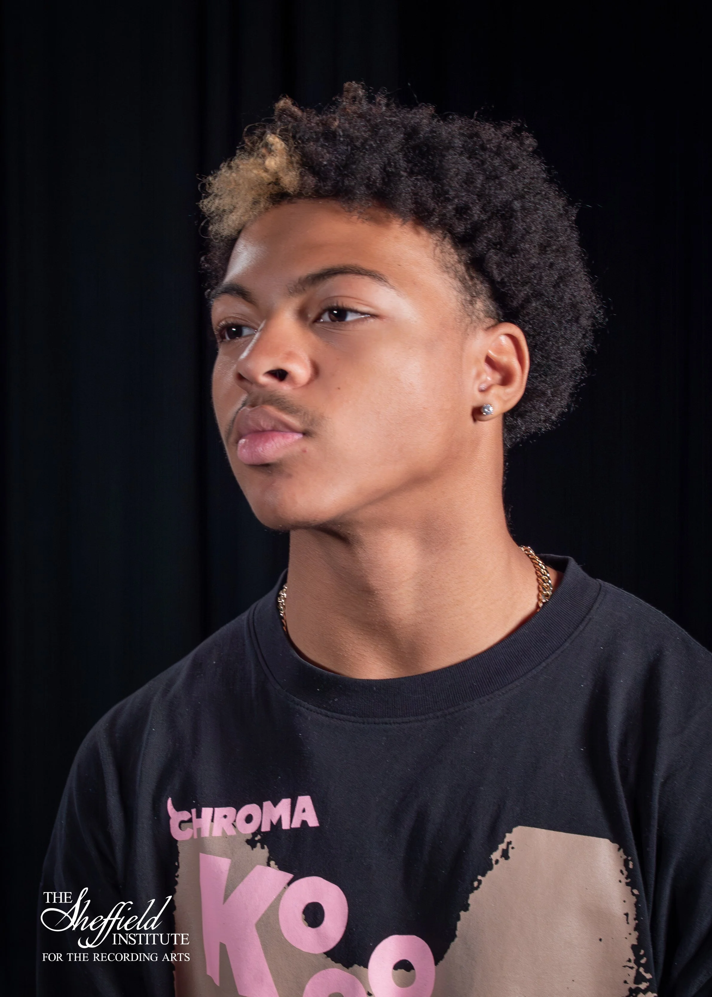 Close-up of a young person with short, curly black hair with blond tips, wearing earrings, a necklace, and a black T-shirt with pink and white text, standing against a dark background.