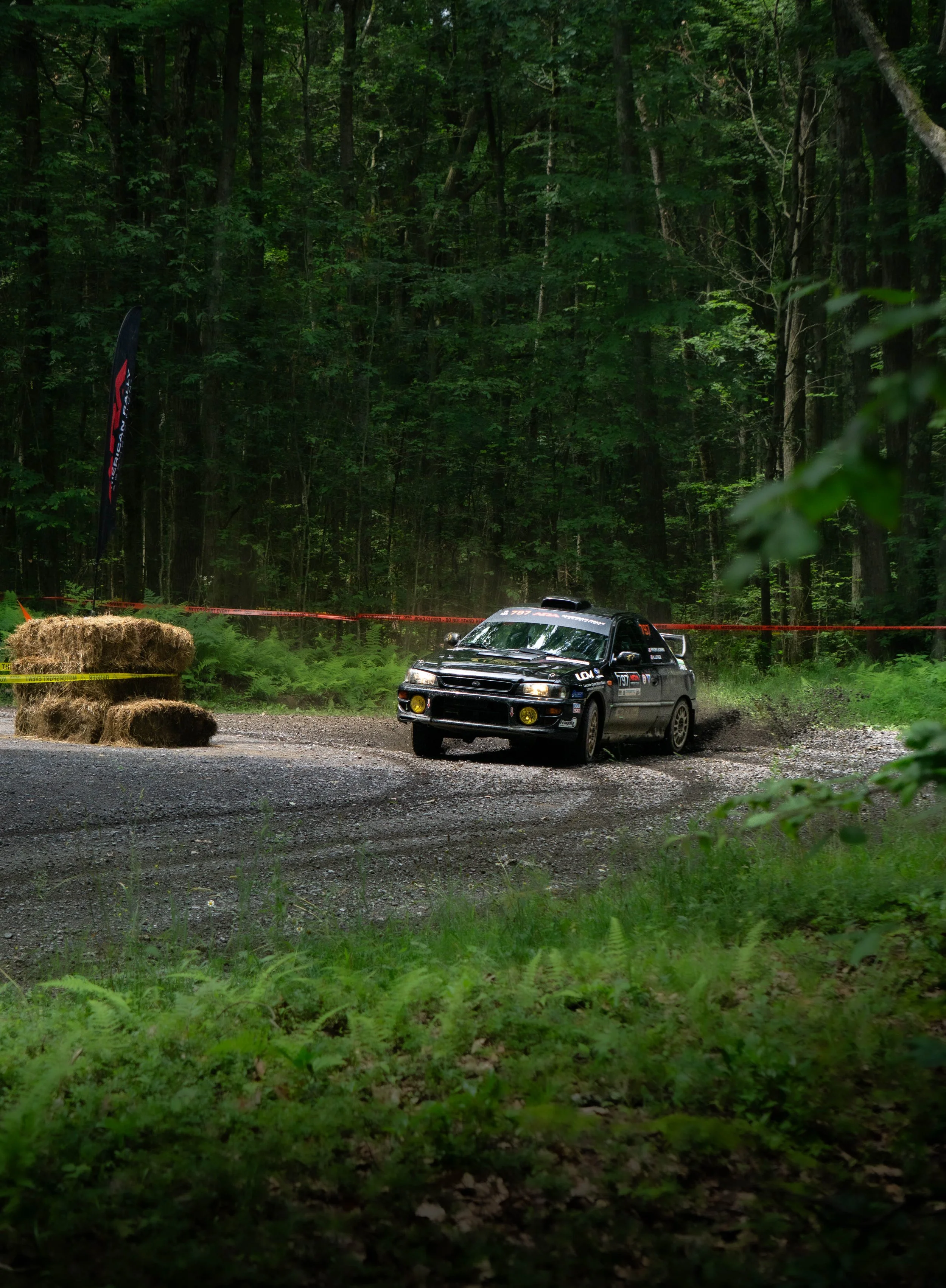 A black rally car with yellow headlights and various sponsor decals is driving on a gravel path through a dense green forest, with hay bales on the side and red and yellow caution tape marking the course.