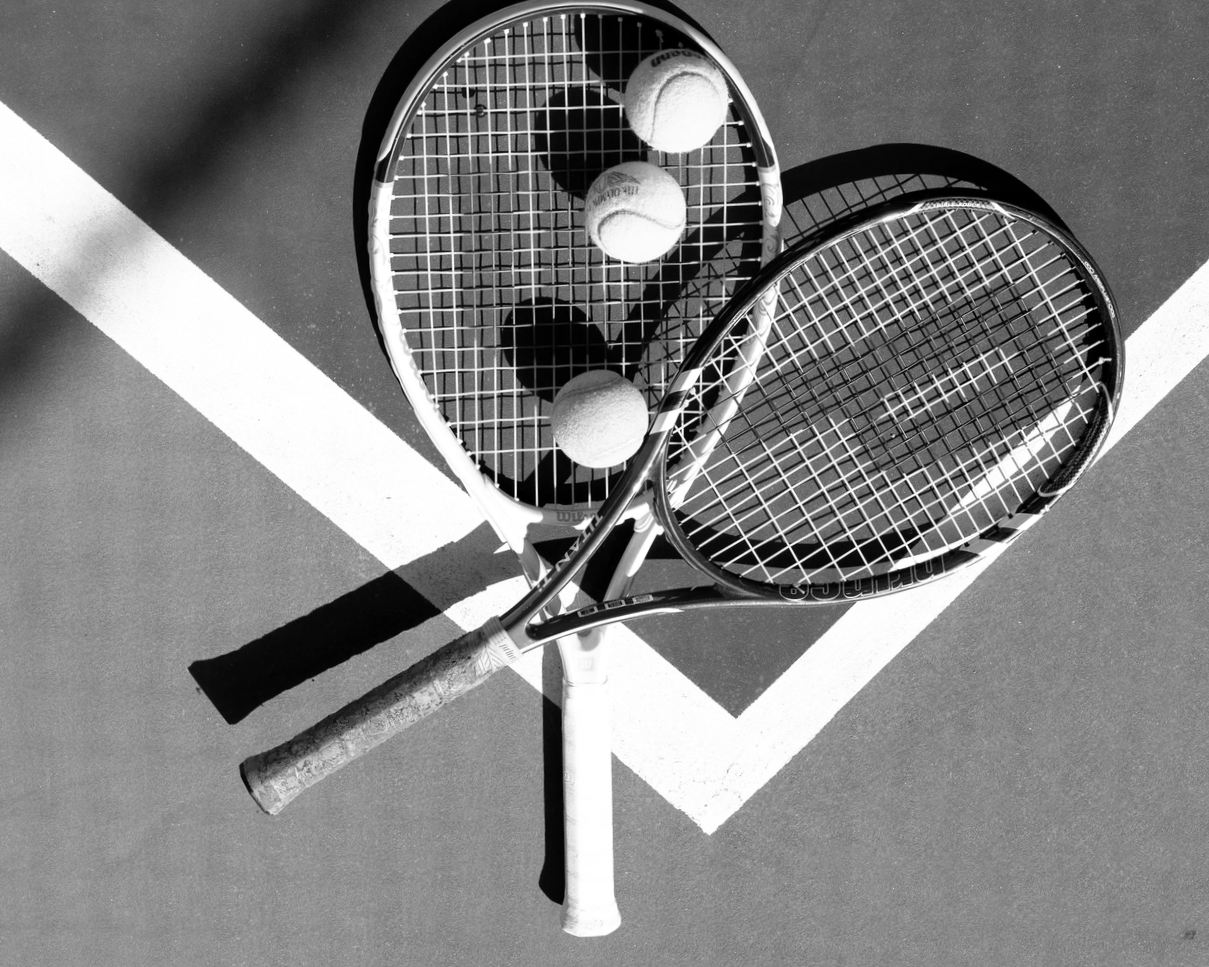 Two tennis rackets crossed over each other on a tennis court with two tennis balls on one racket, in black and white.