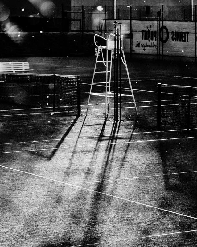 Lighted tennis court at night with a chair, a ball hopper, and court net shadows.