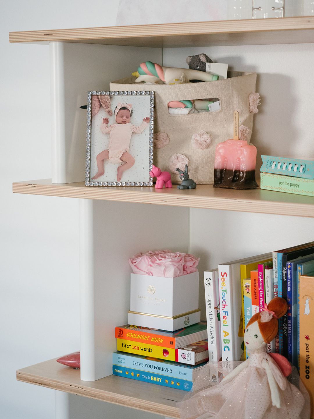 Bedside shelf with framed baby photo, plush toys, a candle-shaped lamp, children's books, a decorative box of pink tissue paper, and a fabric doll with red hair.