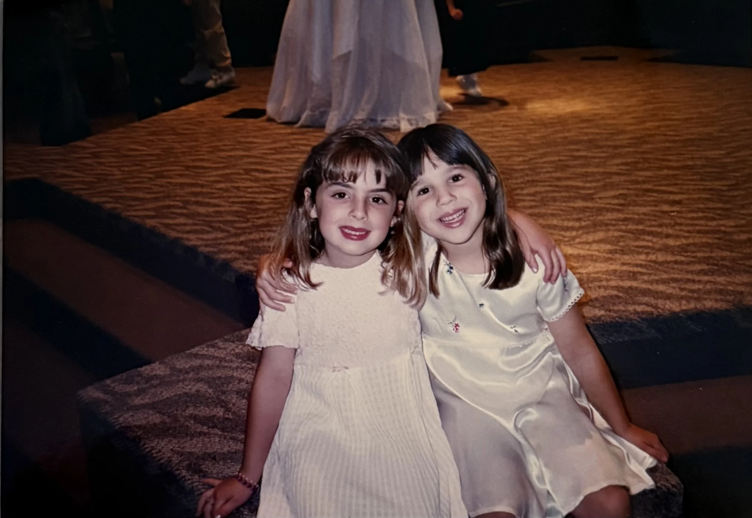 Two young girls sitting on the floor with their arms around each other, smiling at the camera. They are wearing light-colored dresses, and there is a carpeted and stage-like area in the background.
