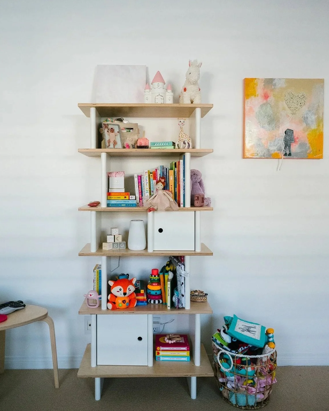 A wooden bookshelf with multiple shelves, decorated with toys, books, and art, standing against a white wall.