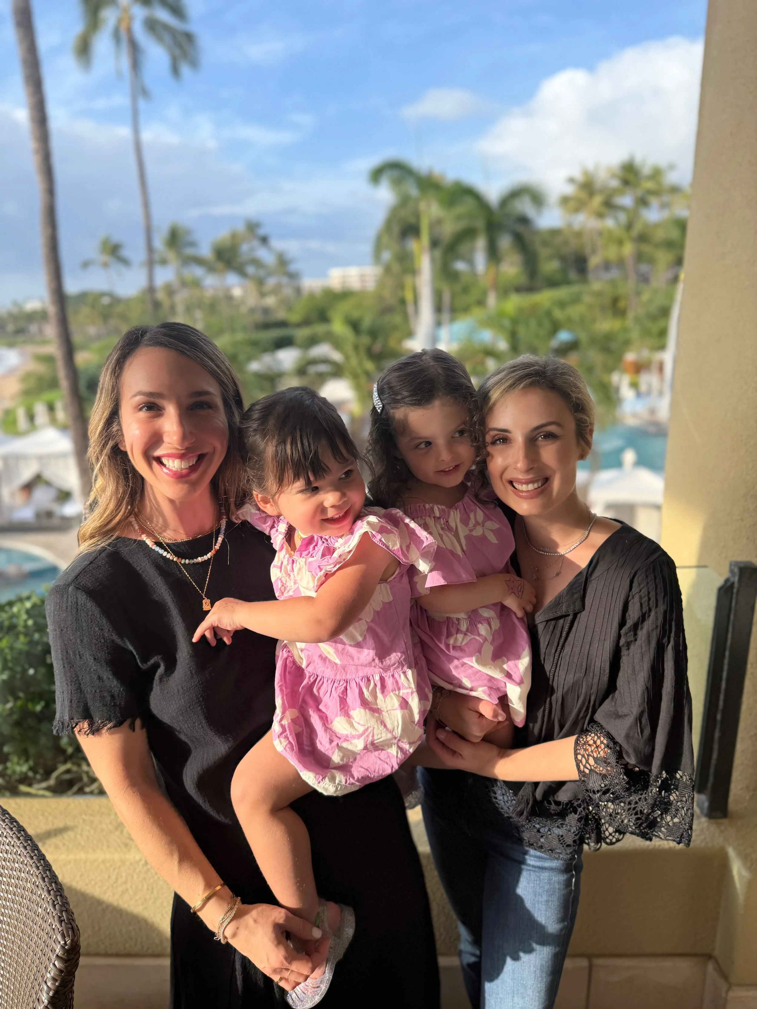 Four women, two adults and two children, smiling and posing together on a balcony with palm trees and a pool in the background, during daytime.