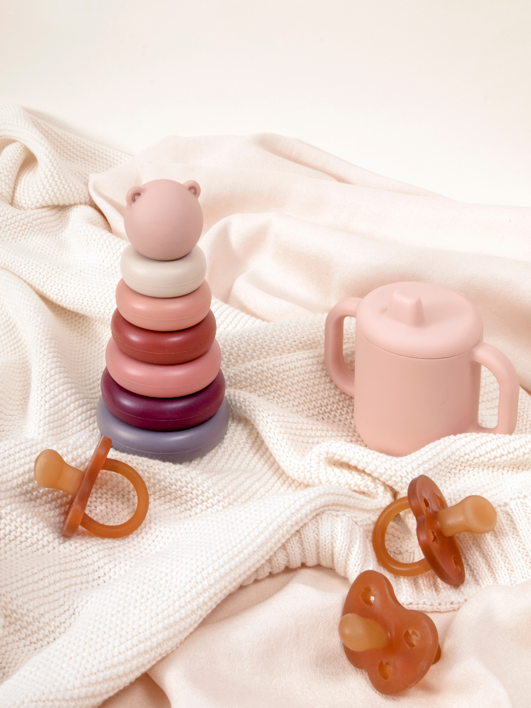 Stack of pastel-colored teething rings with a teddy bear shape on top, pink sippy cup with handles, and a pacifier, all arranged on cream-colored textured fabrics.