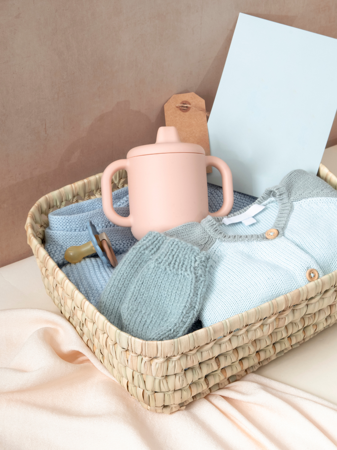 A basket filled with baby items including a pink sippy cup, a blue baby blanket, a light blue baby cardigan with buttons, a baby comb, and a pacifier, placed on a pink satin fabric against a light brown background with a white card and a brown tag.