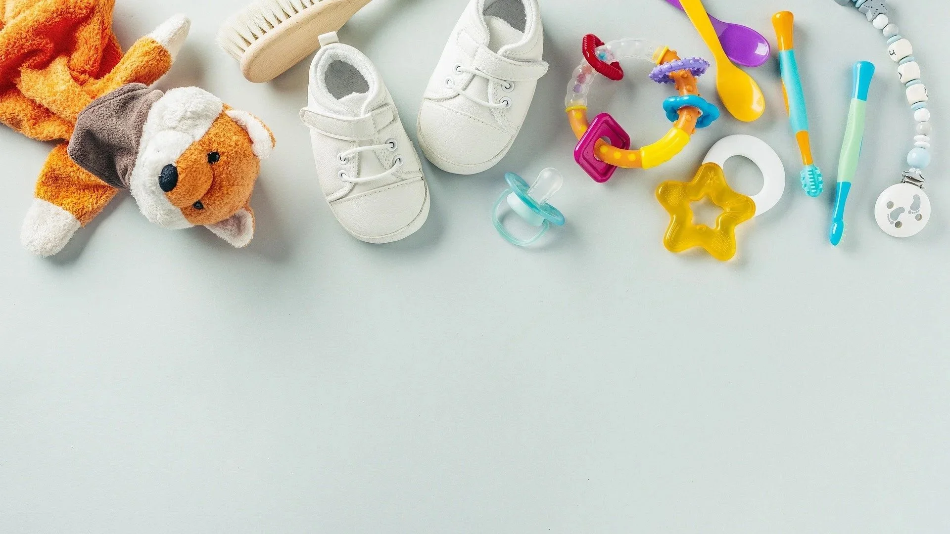 Baby toys including plush dog, baby shoes, pacifiers, colorful teething rings, spoons, and toothbrushes on a light background.