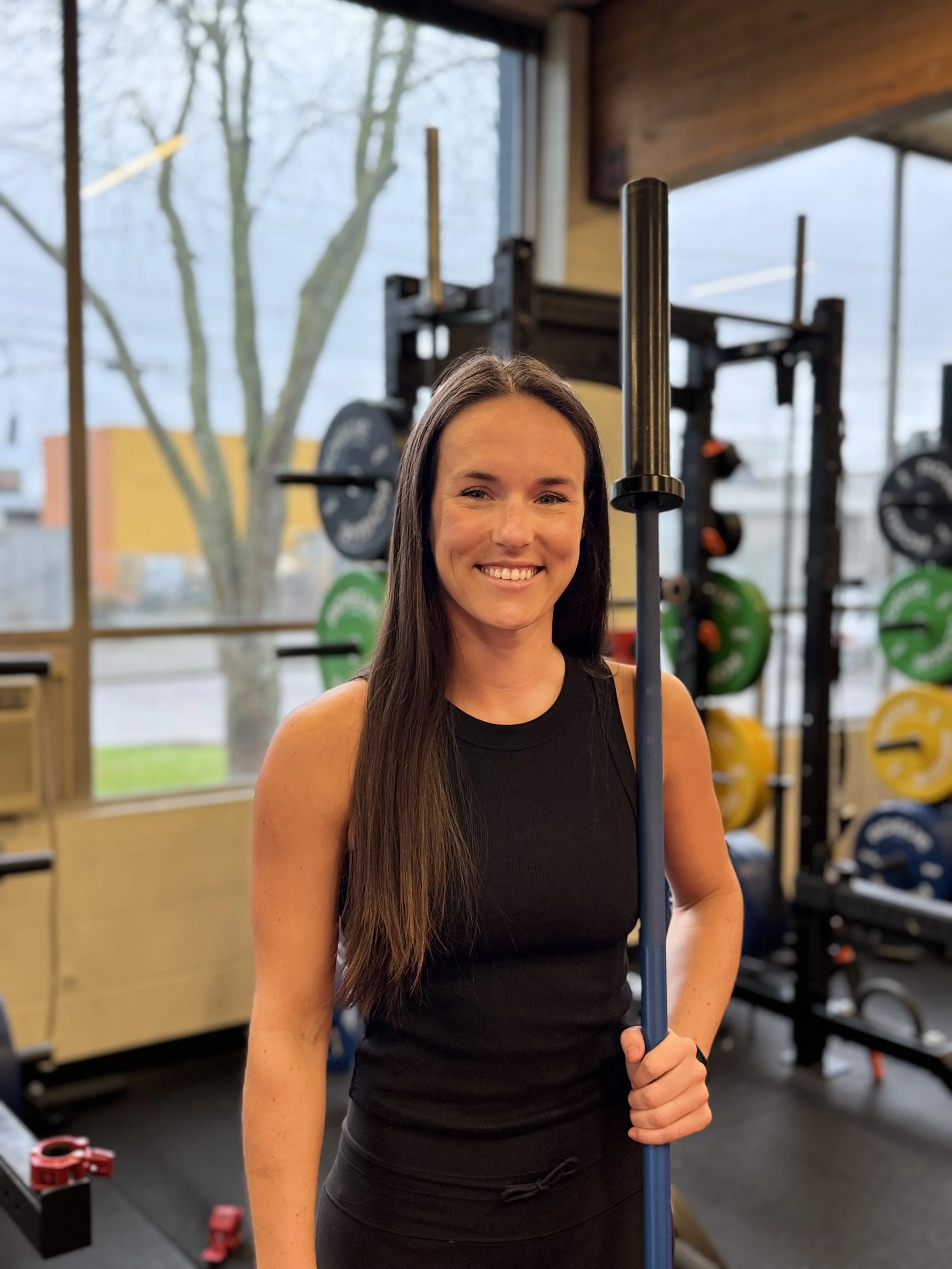A woman with long brown hair, wearing a black sleeveless top, smiling, holding a blue barbell inside a gym with weightlifting equipment in the background.