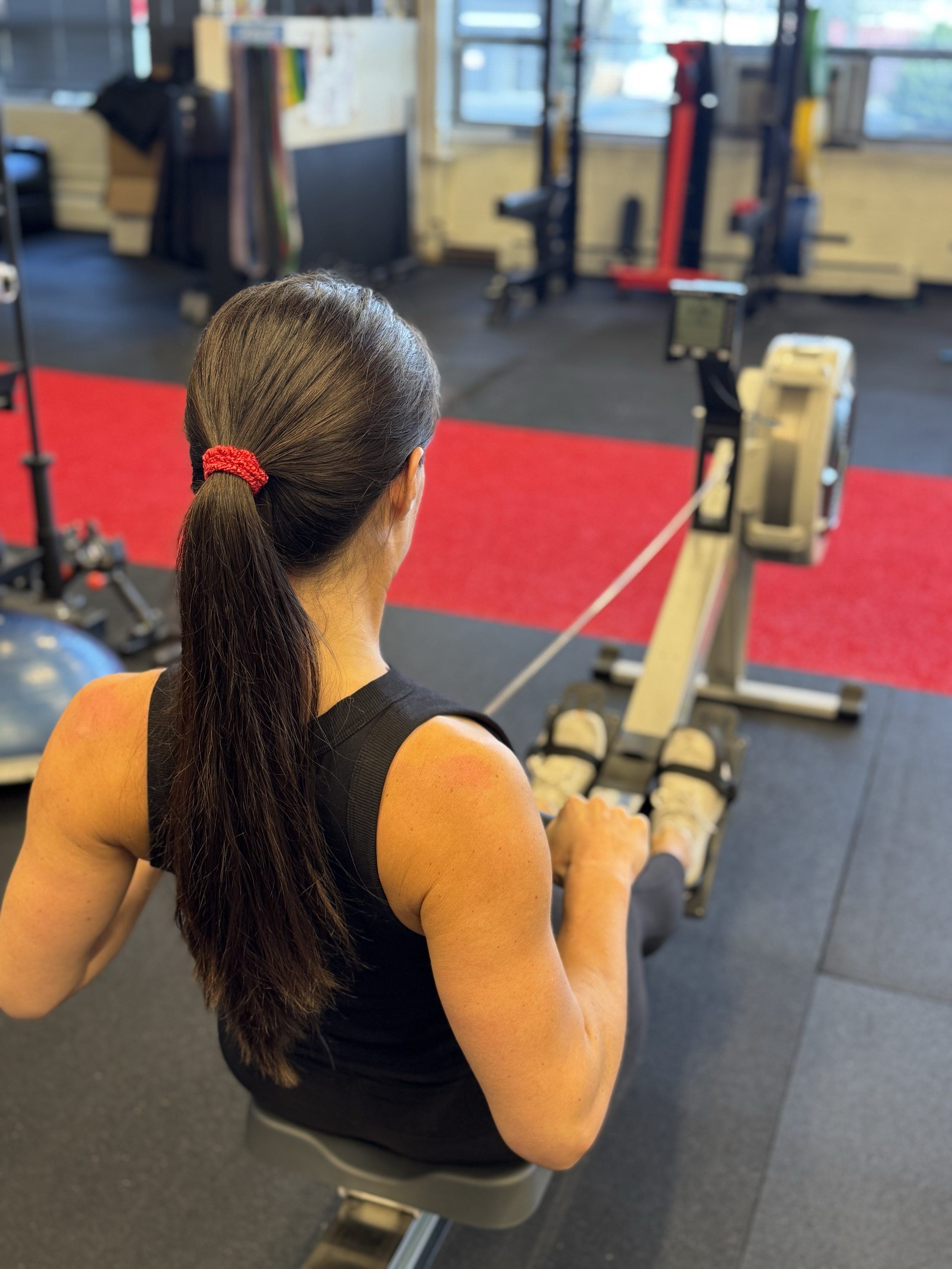 Woman using a rowing machine at a gym, viewed from behind, with workout equipment and windows in the background.