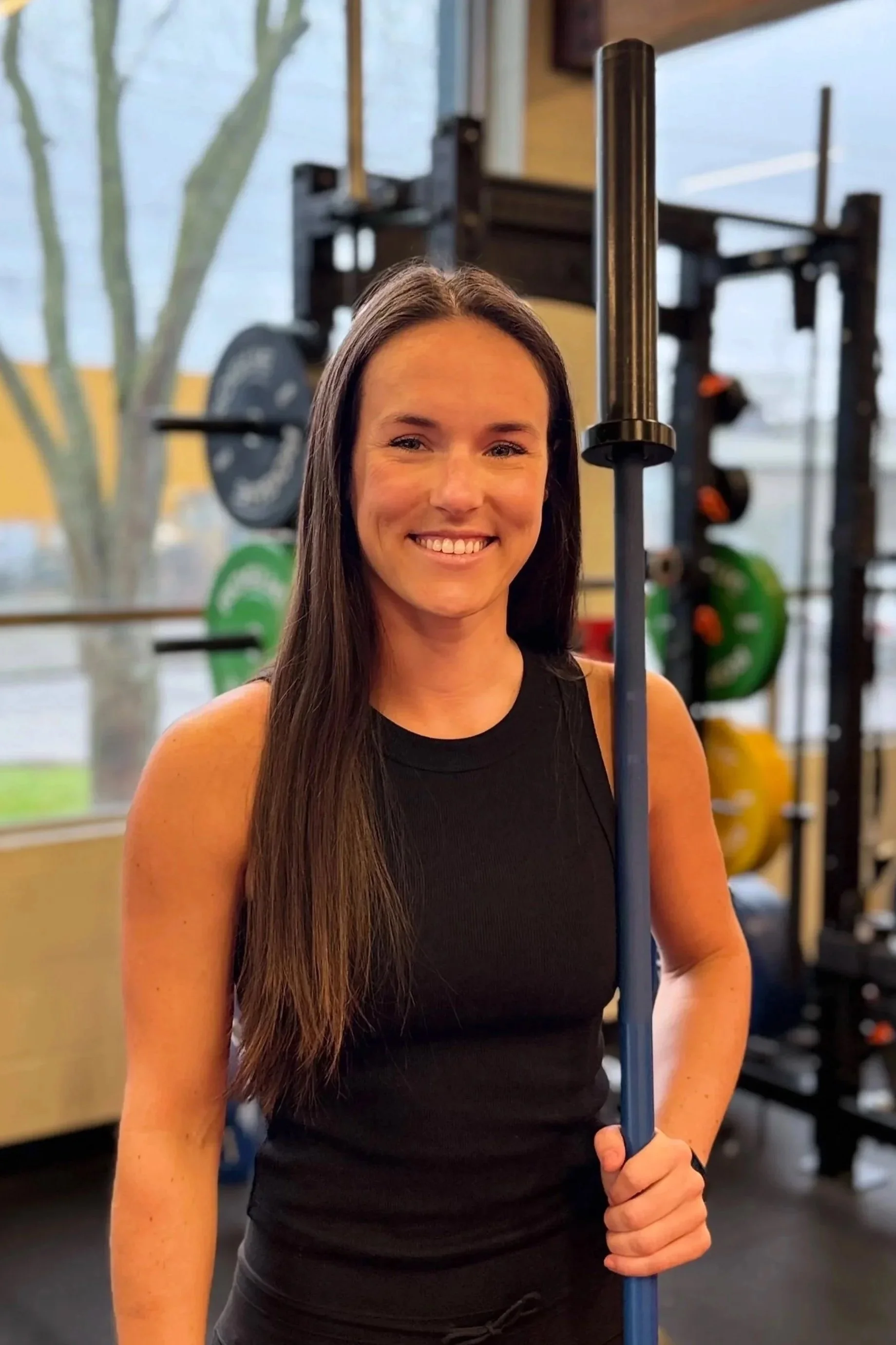 A woman smiling at the gym, holding a barbell stake, with weightlifting equipment in the background.