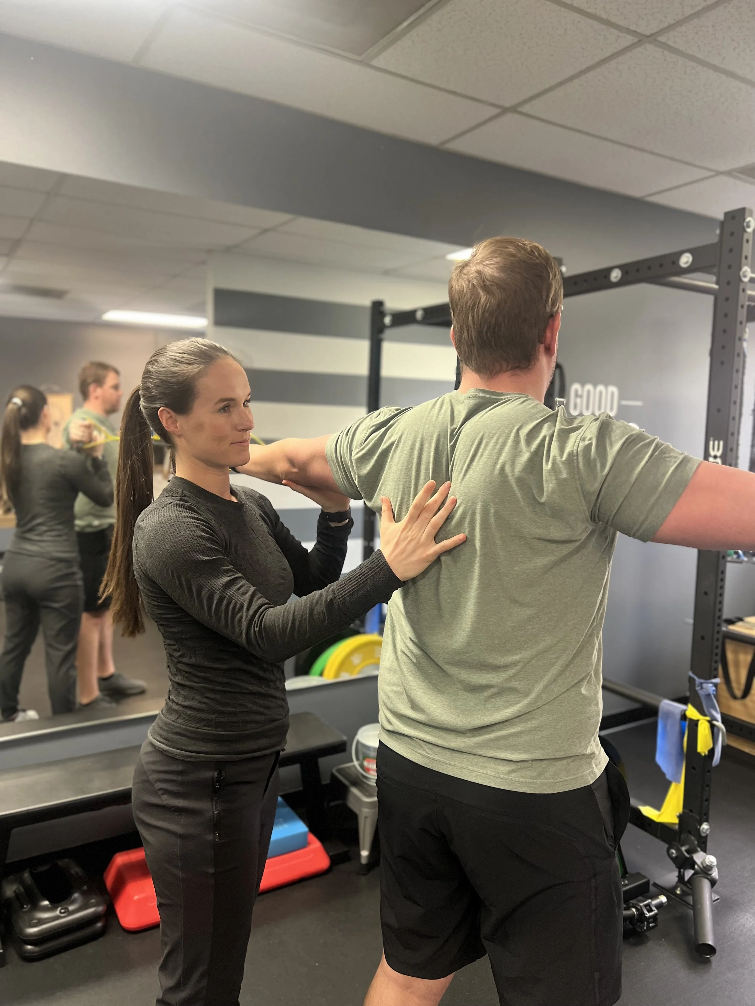 A woman assisting a man with shoulder stretching in a gym.