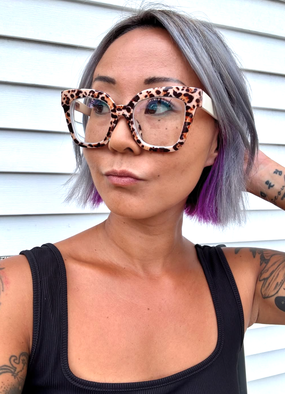A woman with shoulder-length gray and purple hair wearing large leopard-print glasses and a black tank top, standing outside in front of white siding.