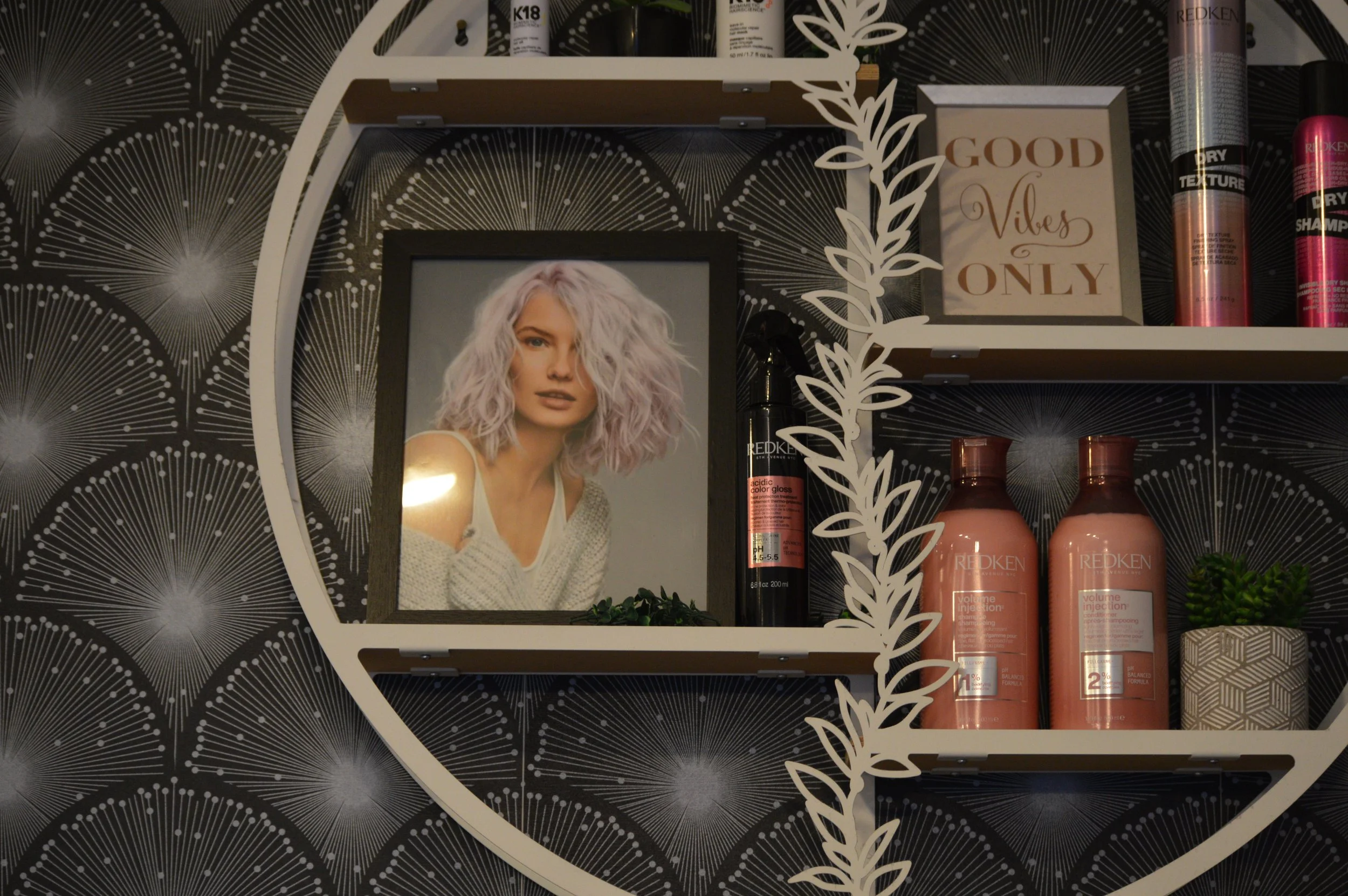A salon shelf with hair care products, a framed photograph of a woman with curly purple hair, and a sign that says 'Good Vibes Only' against black wallpaper with white radial patterns.
