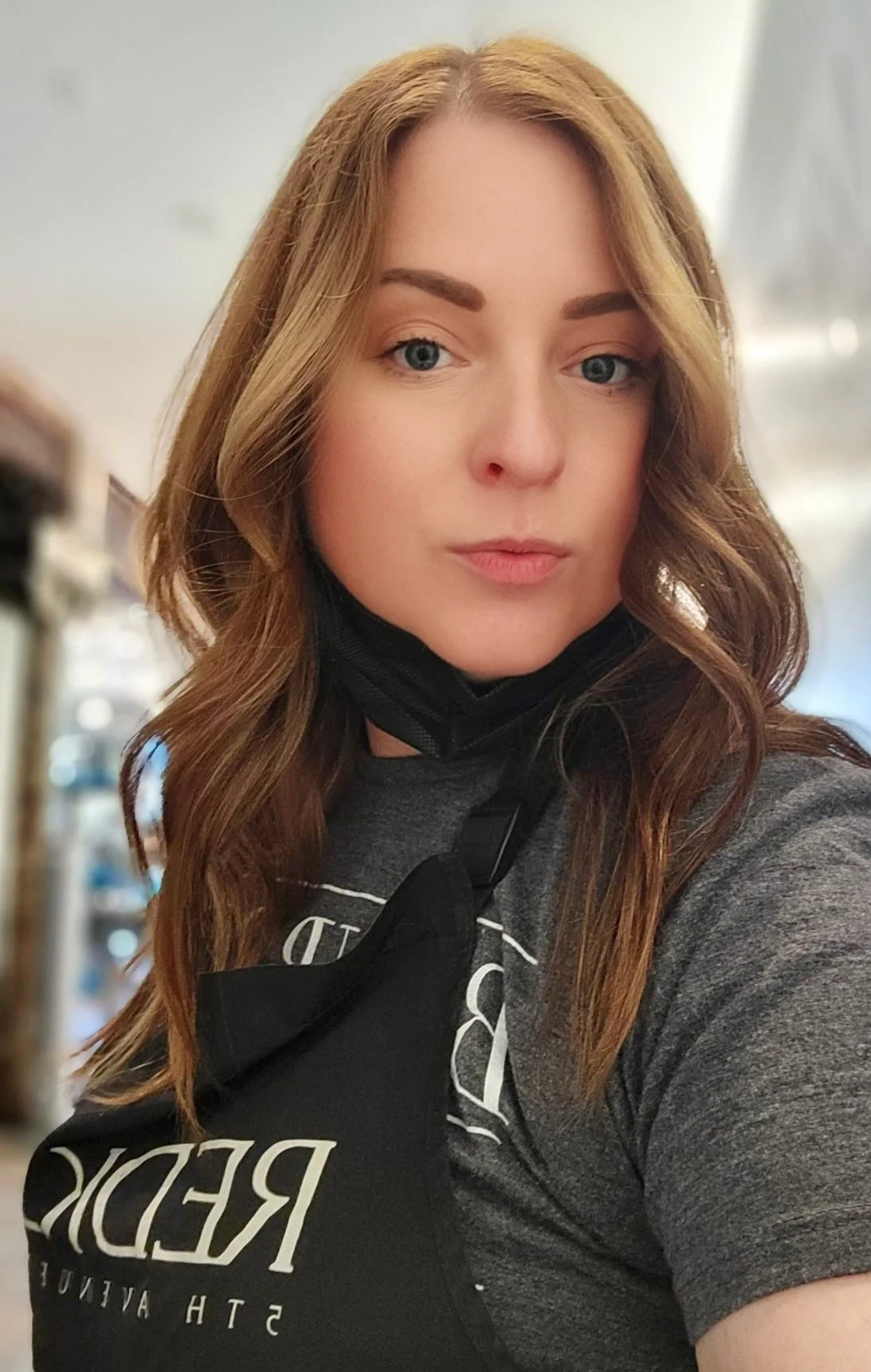 A woman with long red hair, blue eyes, and makeup, wearing a gray shirt and a black apron, taking a selfie in what appears to be a store or café.