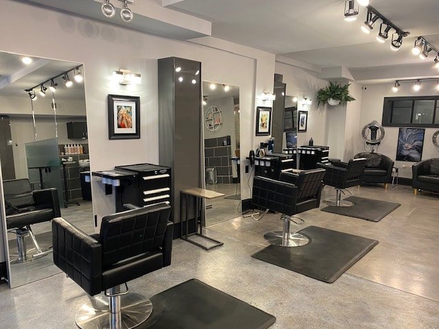 Empty hair salon with four styling stations, black chairs, large mirrors, and salon equipment, with framed pictures on the walls and a ceiling light fixture.