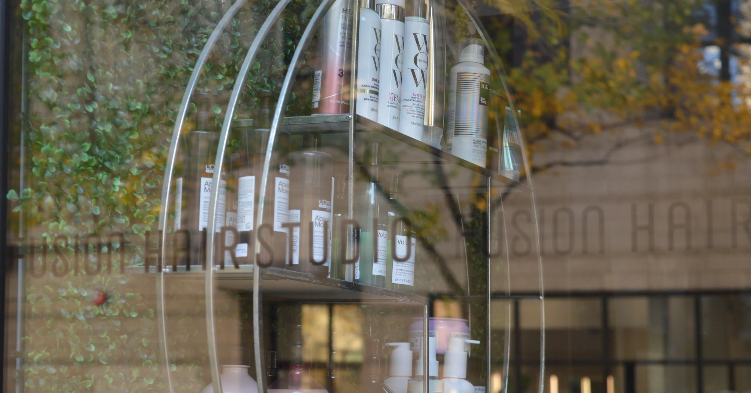 Glass window showing shelves with hair care products, including sprays and shampoos, in a salon or store, with trees and a building visible outside.