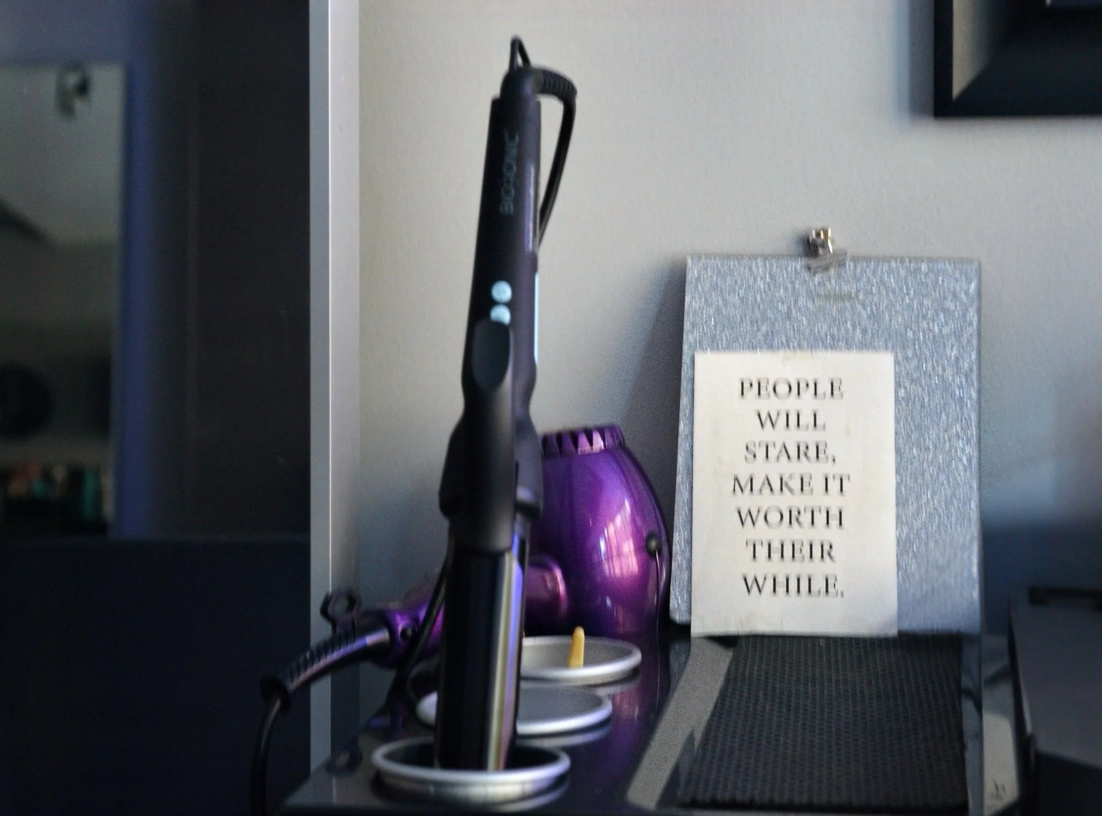 Desk with a purple vacuum, a black electric toothbrush, a tealight candle holder, a glittery box with a motivational quote, and a black mat.