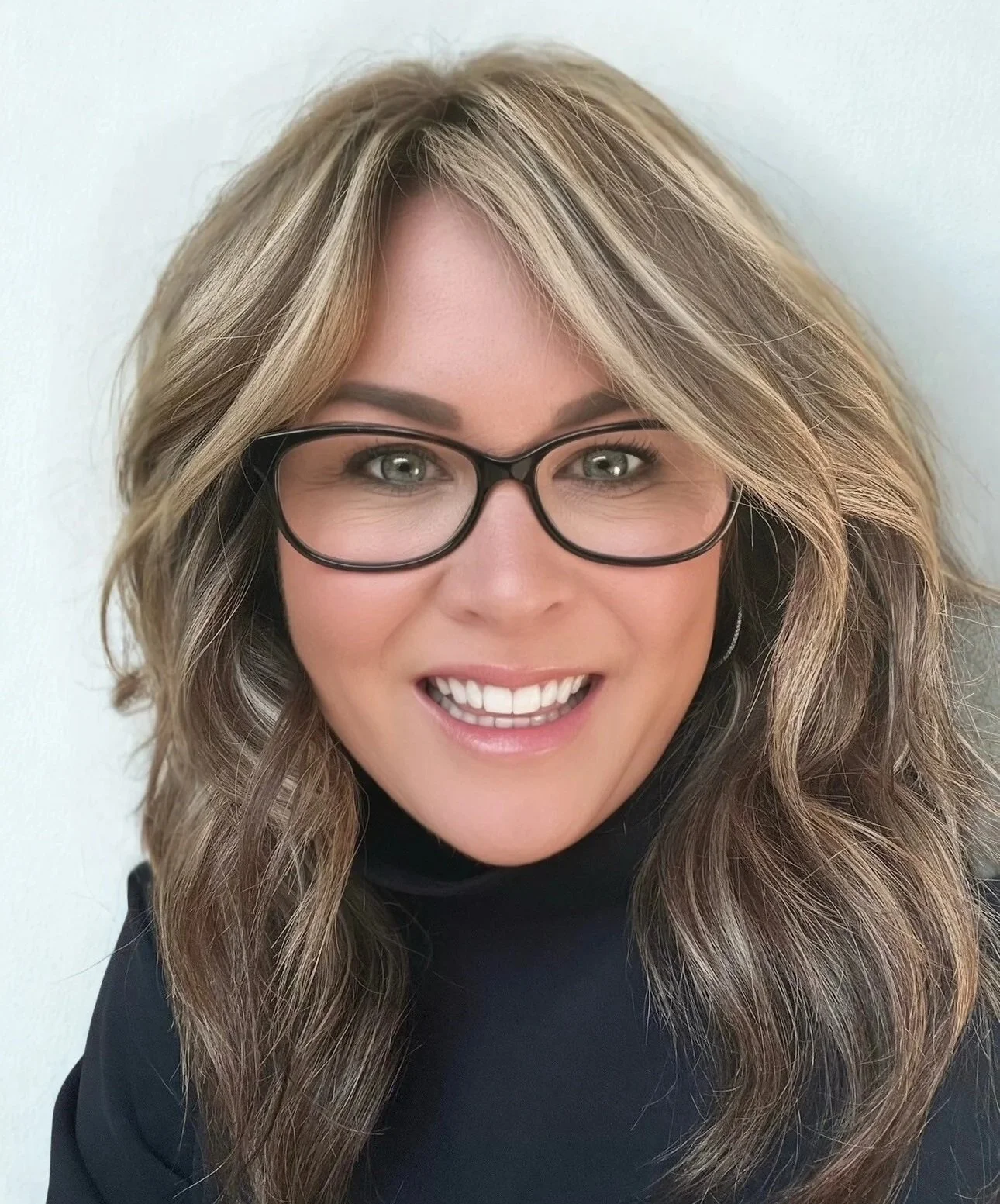 A woman with wavy light brown hair, glasses, and a smile, wearing a black top.