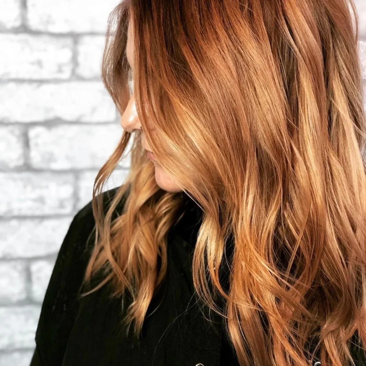 Close-up of a woman with wavy, reddish-brown hair against a white brick wall background.