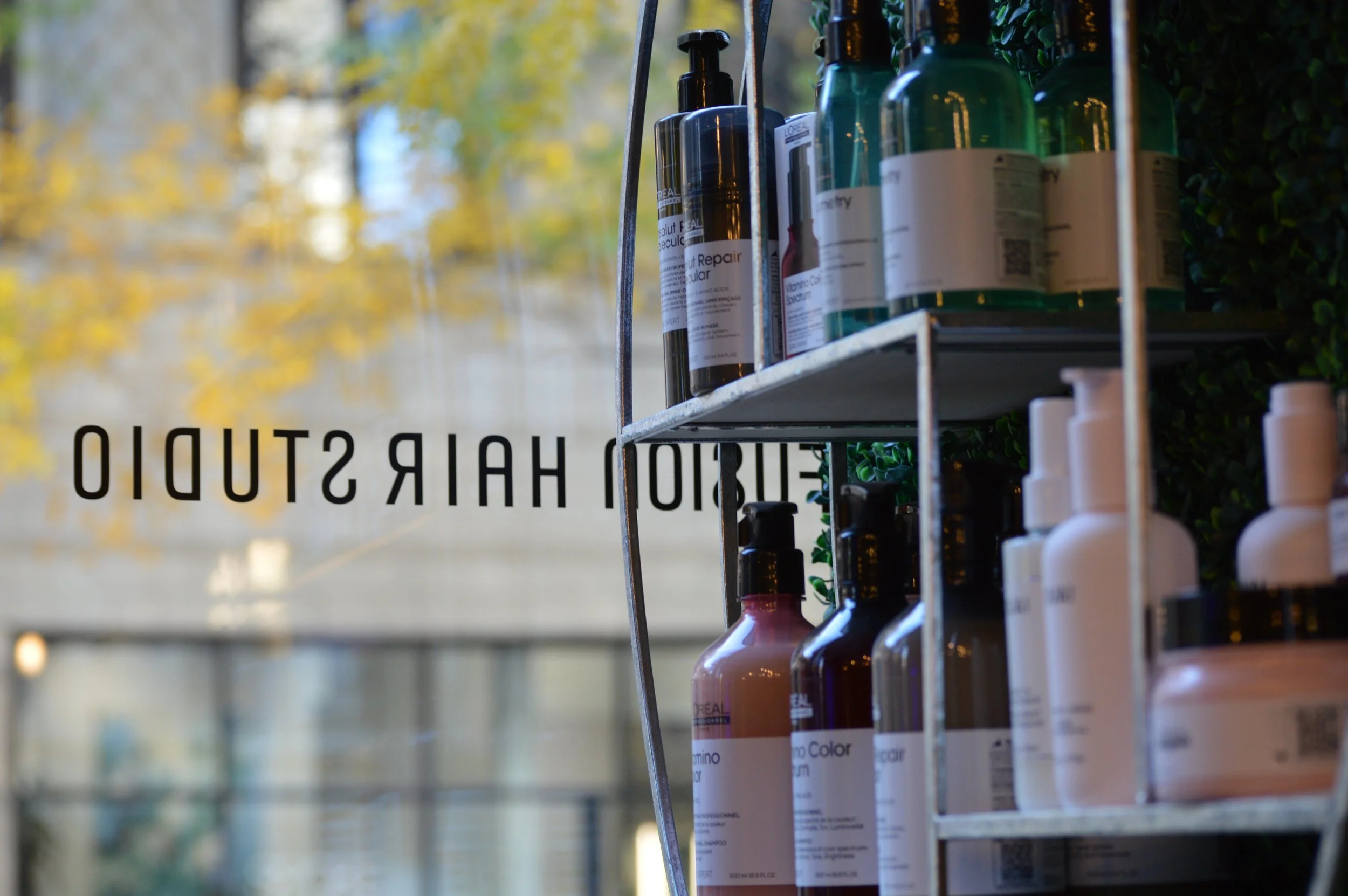 Shelves with various hair care and styling products, including bottles and pump dispensers, inside a salon or store with a glass window reflecting outside trees.