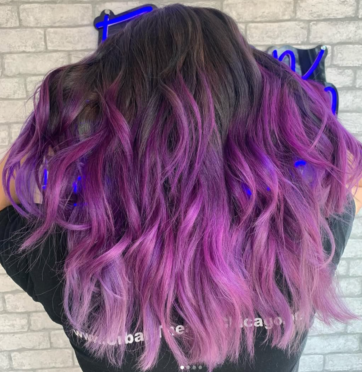 Person with long wavy hair dyed in shades of purple and pink, standing in front of a brick wall with neon sign.