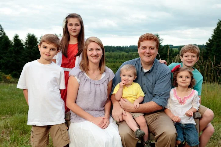Family of seven sitting outdoors in a grassy field with trees in the background, smiling at the camera.