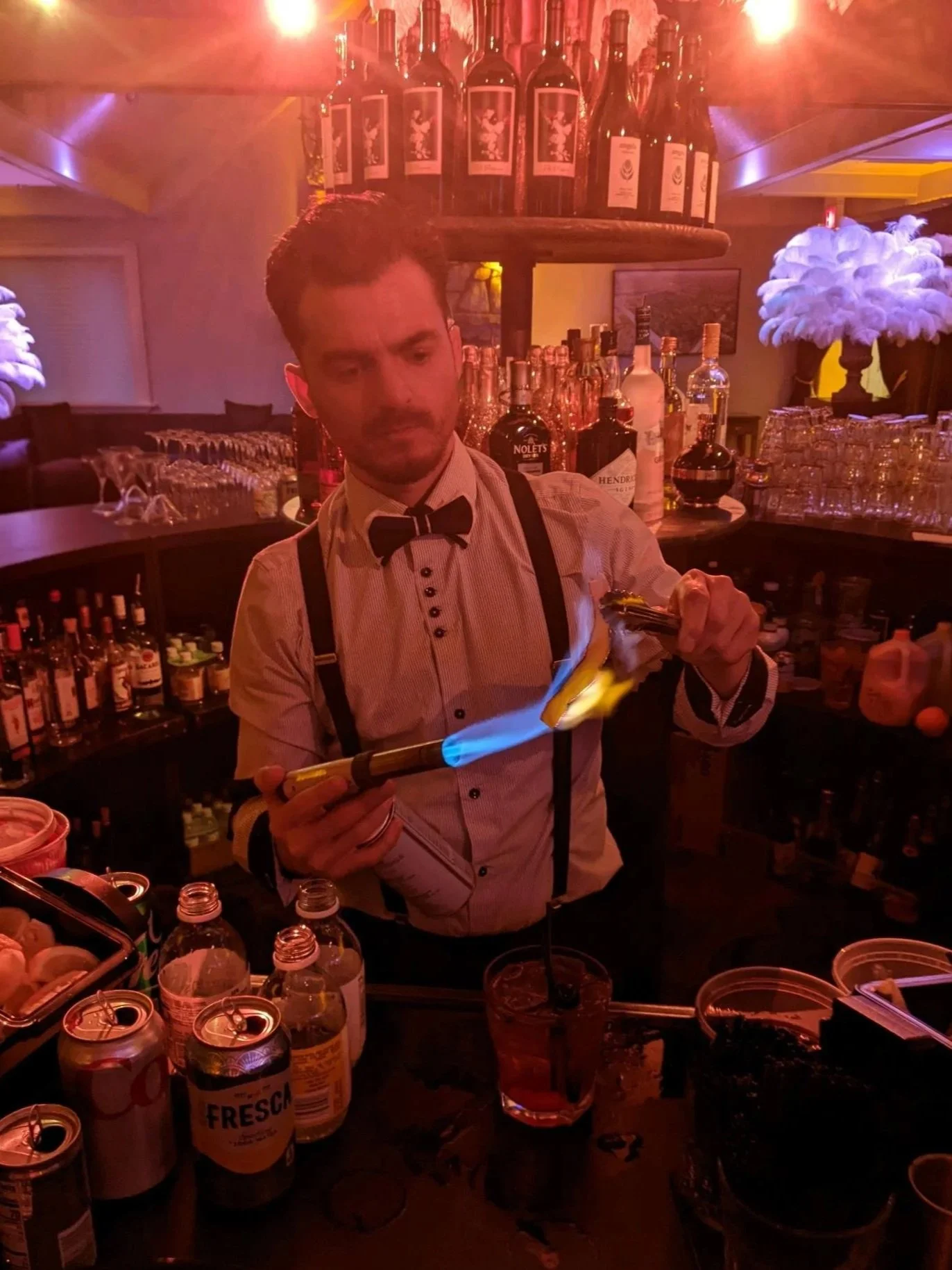 A bartender in a white shirt with suspenders and a bow tie is lighting a drink on fire at a bar with bottles of alcohol and glasses in the background.