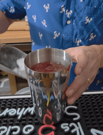 Person pouring a red beverage into a stainless steel cup on a bar counter.