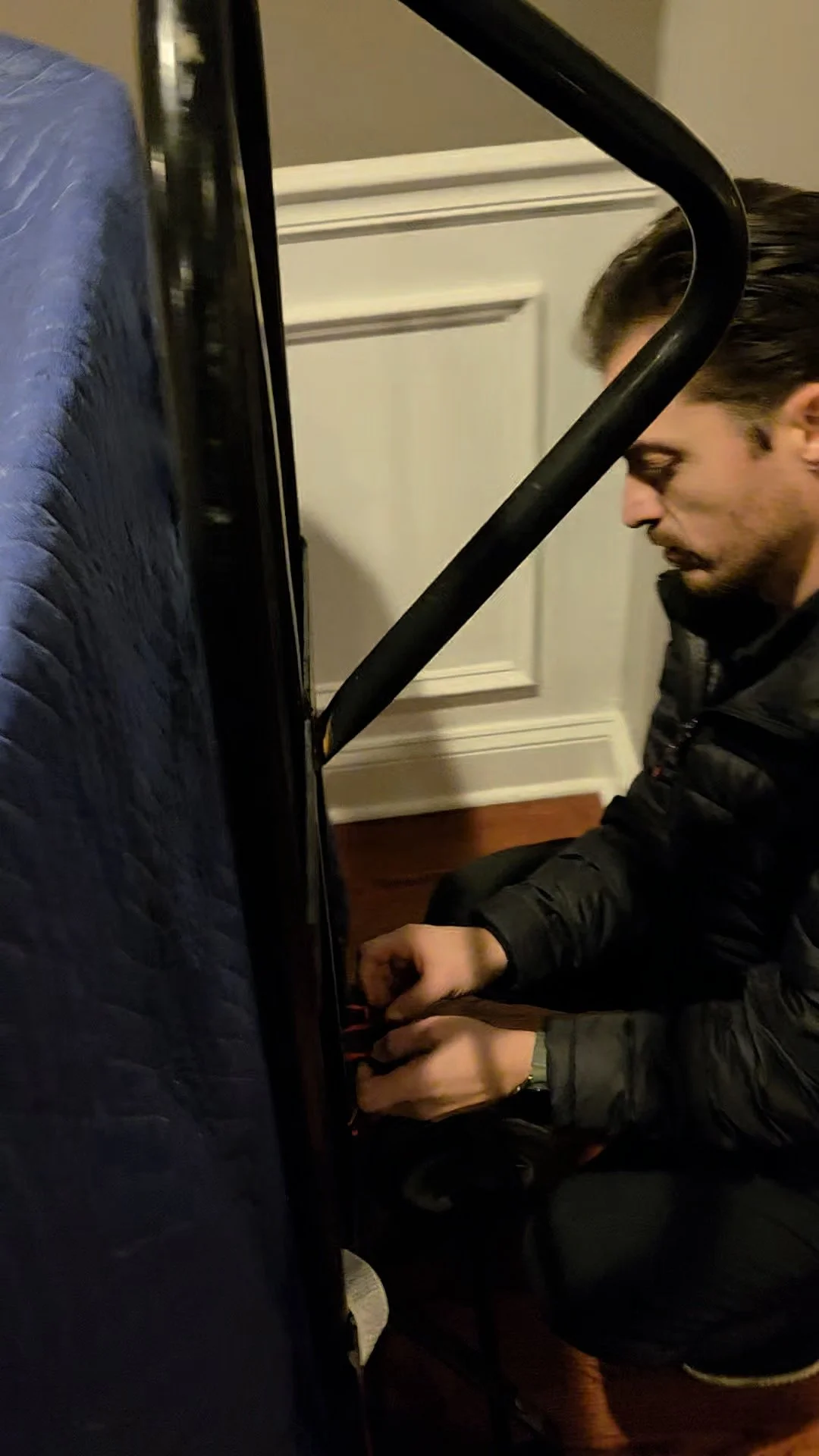 A man dressed in a black jacket and black pants kneeling beside a black piano, with his hands on the keyboard, indoors with a white door and wooden floor in the background.