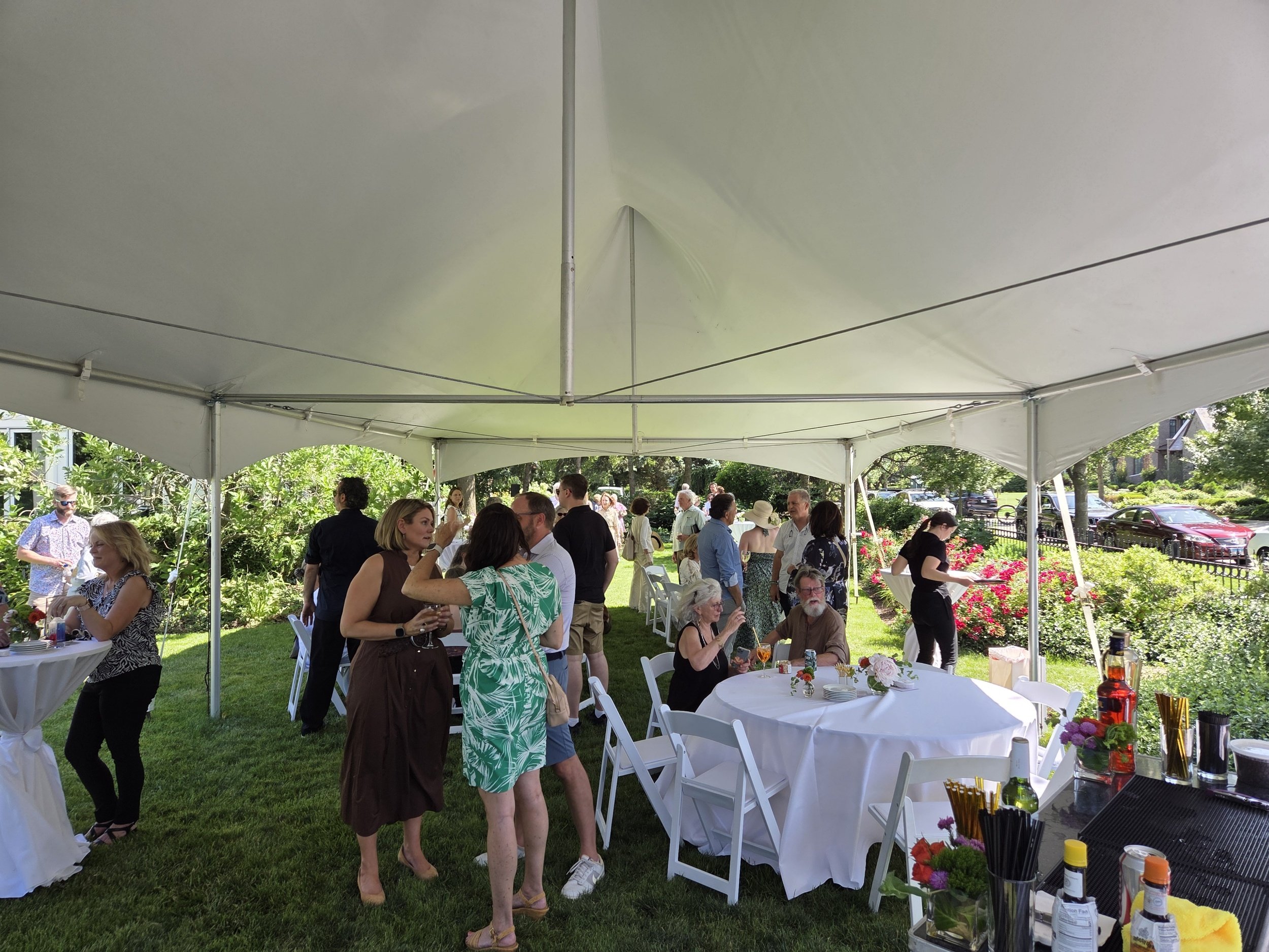 People gathered under a large white tent at an outdoor event in a garden with green trees and bushes, some sitting at round tables with white tablecloths, others standing and talking, with food, drinks, and flowers on the tables.