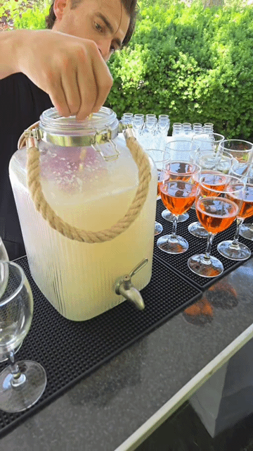 Person pouring lemonade from a large glass dispenser with a rope handle into a glass on an outdoor table.