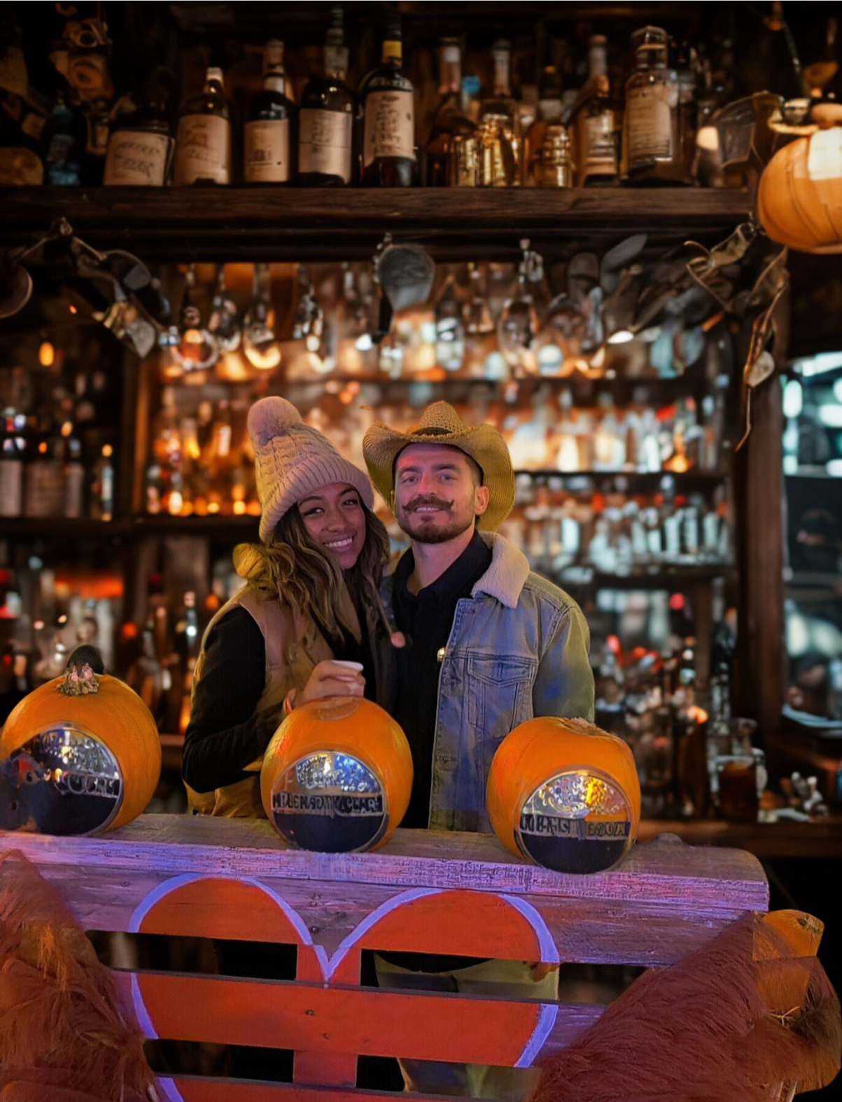 Two people, a woman and a man, smiling and wearing hats, stand behind three small pumpkins on a wooden table in a rustic bar or pub setting decorated for Halloween, with bottles and bar accessories in the background.