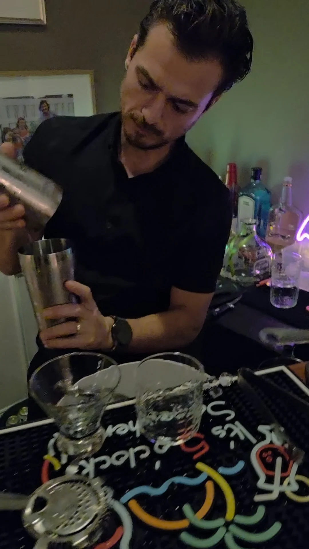 A man in a black shirt preparing a cocktail at a bar with various bottles and drinking glasses in the background.