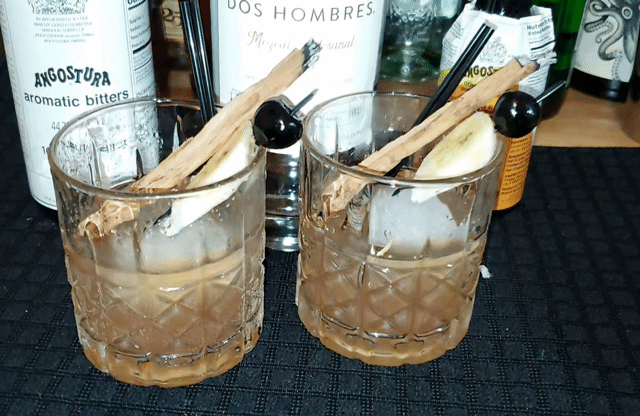Two glasses of dark amber cocktail garnished with banana slices, cinnamon sticks, and black cherries with bottled bitters and liquor bottles in the background.
