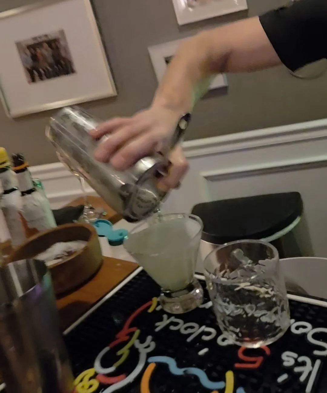 A person pouring a drink from a metal shaker into a cocktail glass filled with a light-colored beverage.