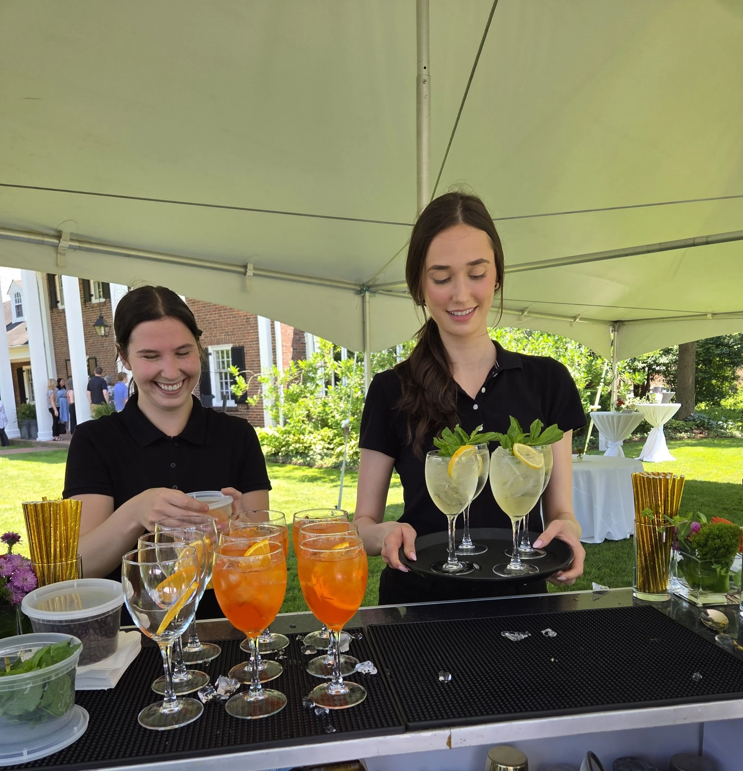 Two smiling women preparing drinks at an outdoor event under a large canopy, with glasses of colorful beverages and lemons on a black serving tray.