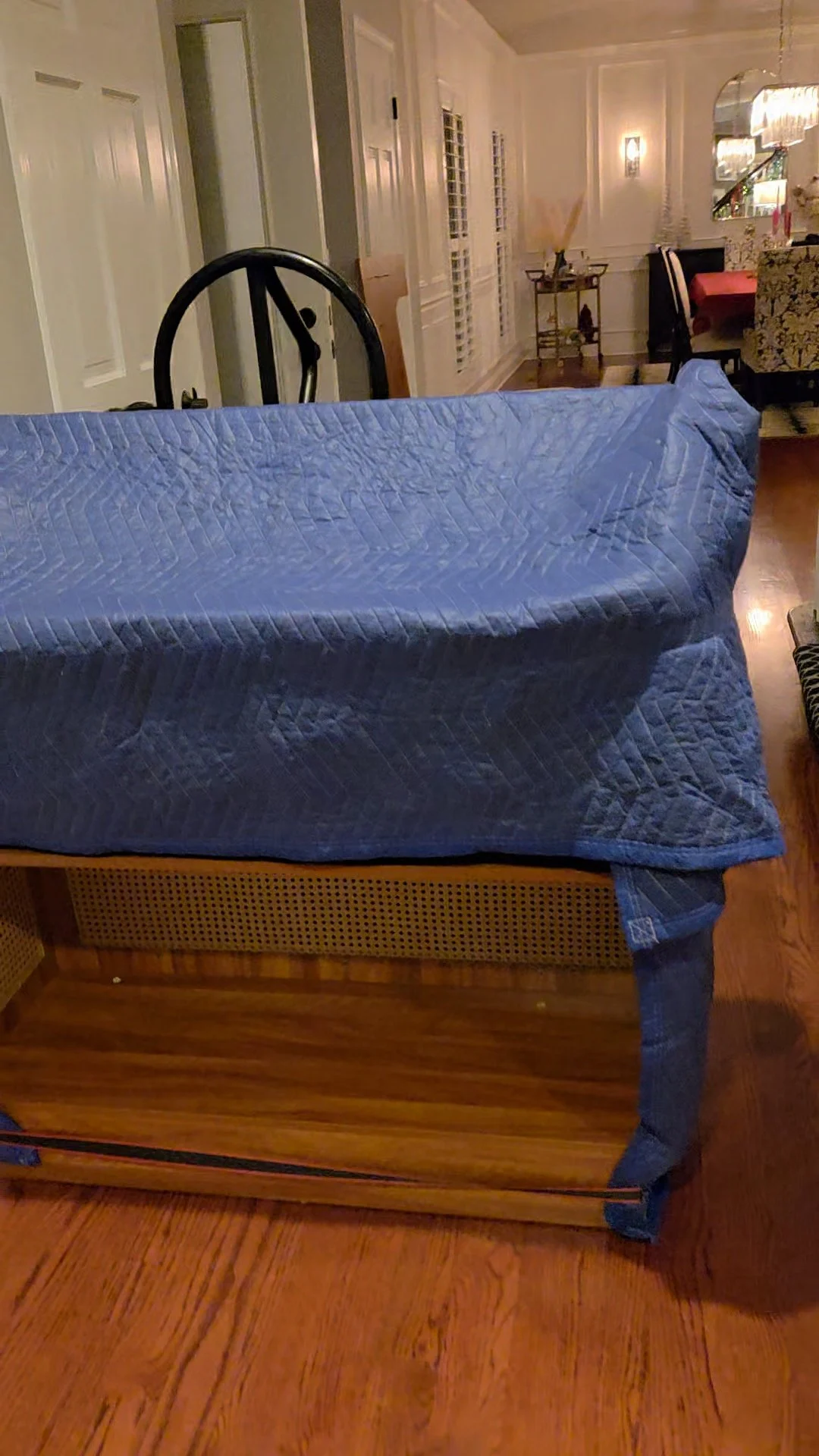 A bed with a blue quilt in a dining room with hardwood floors, black chairs, a chandelier, and a side table with decorative items.